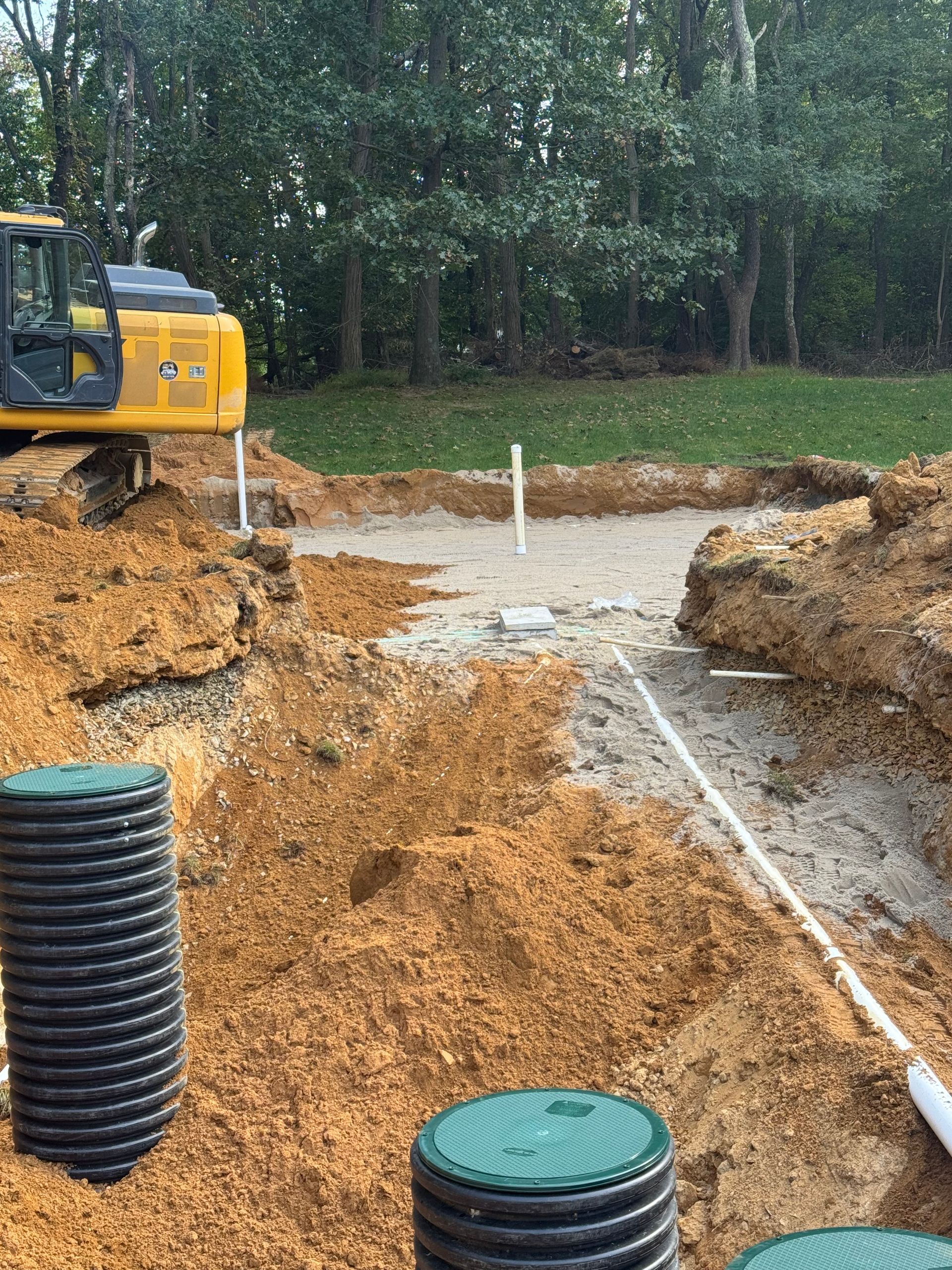 Excavation site with a yellow excavator, plastic pipes, and black drainage tanks; trees in the background.