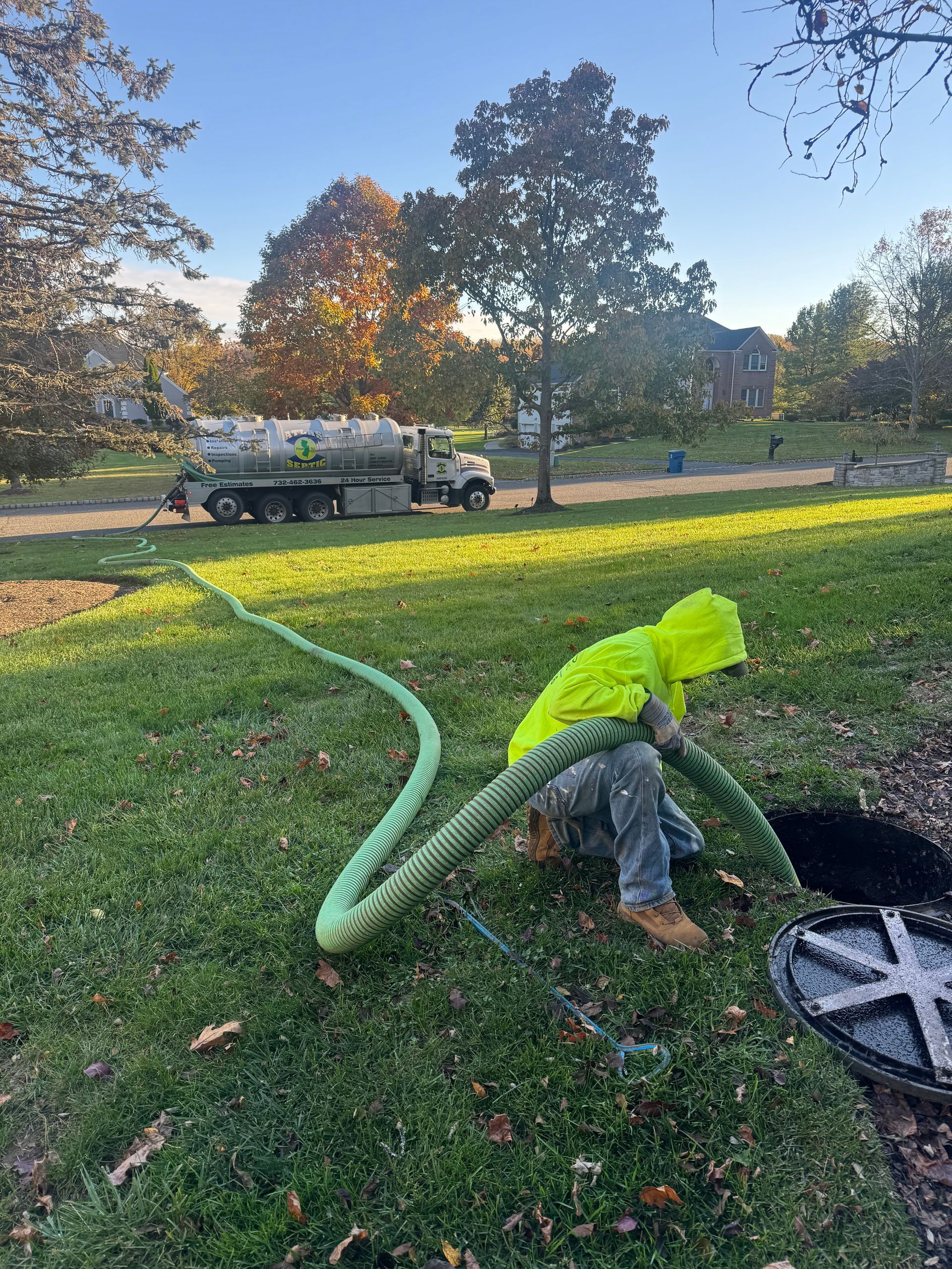 Person in yellow jacket pulling a green rope from an open manhole in a grassy area, truck in background.