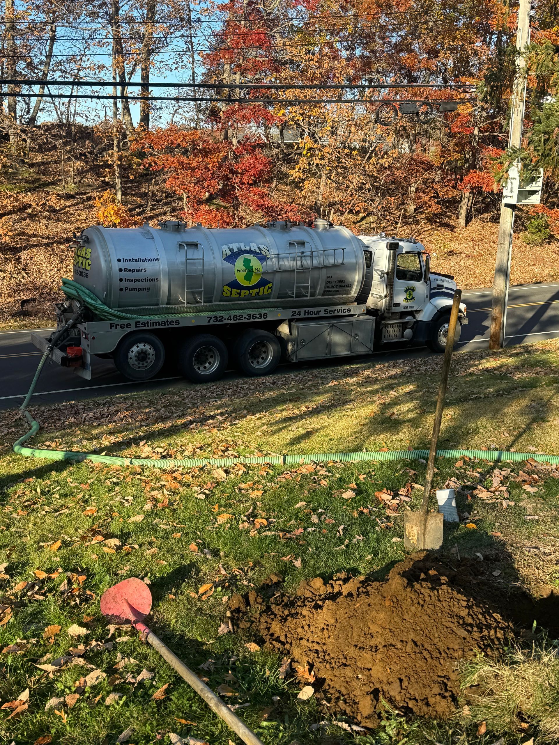 Septic tank truck near a newly planted tree. Shovel in the foreground. Autumn foliage in the background.