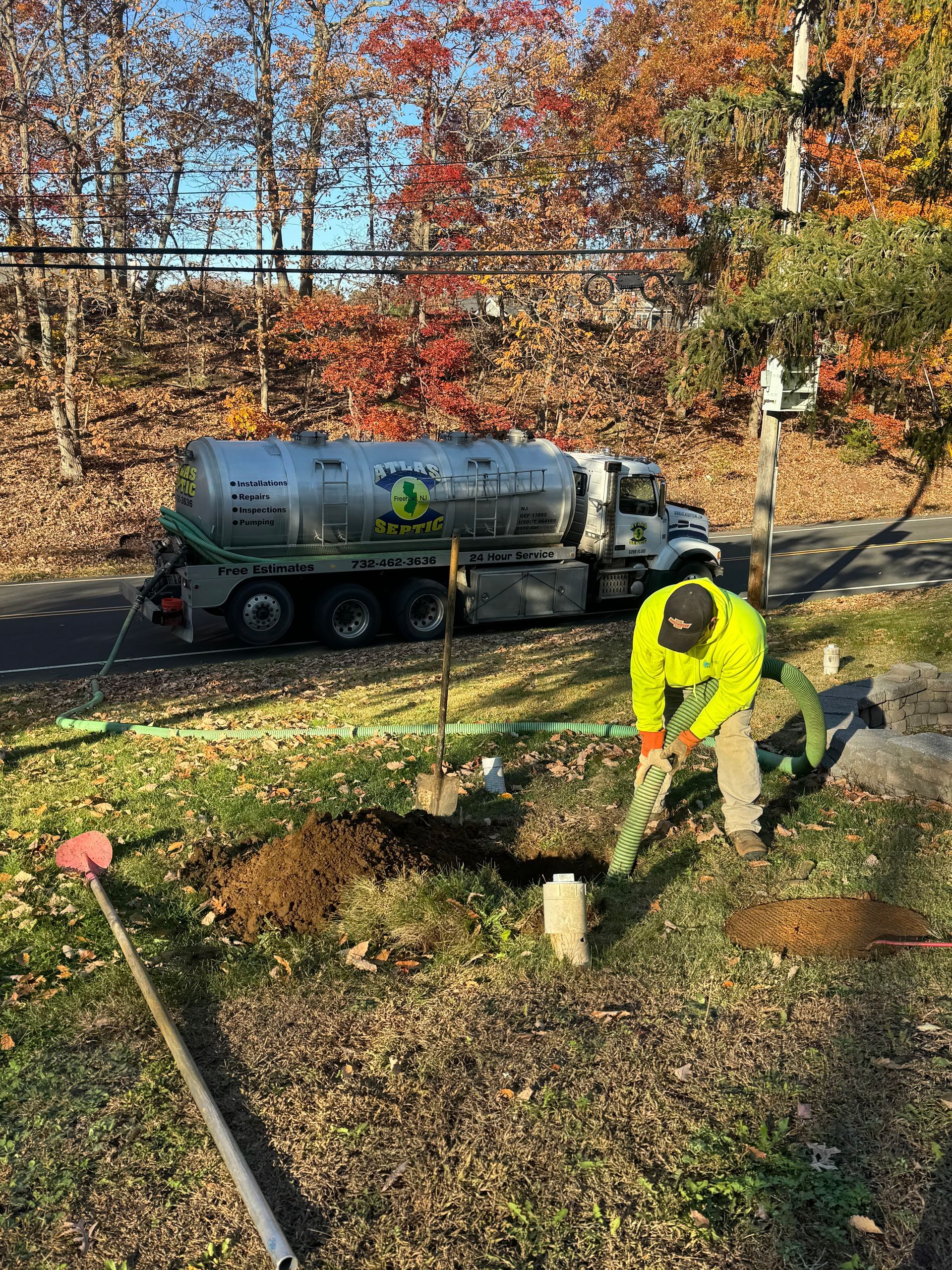Worker digging with a shovel near a septic truck on a roadside, fall foliage.