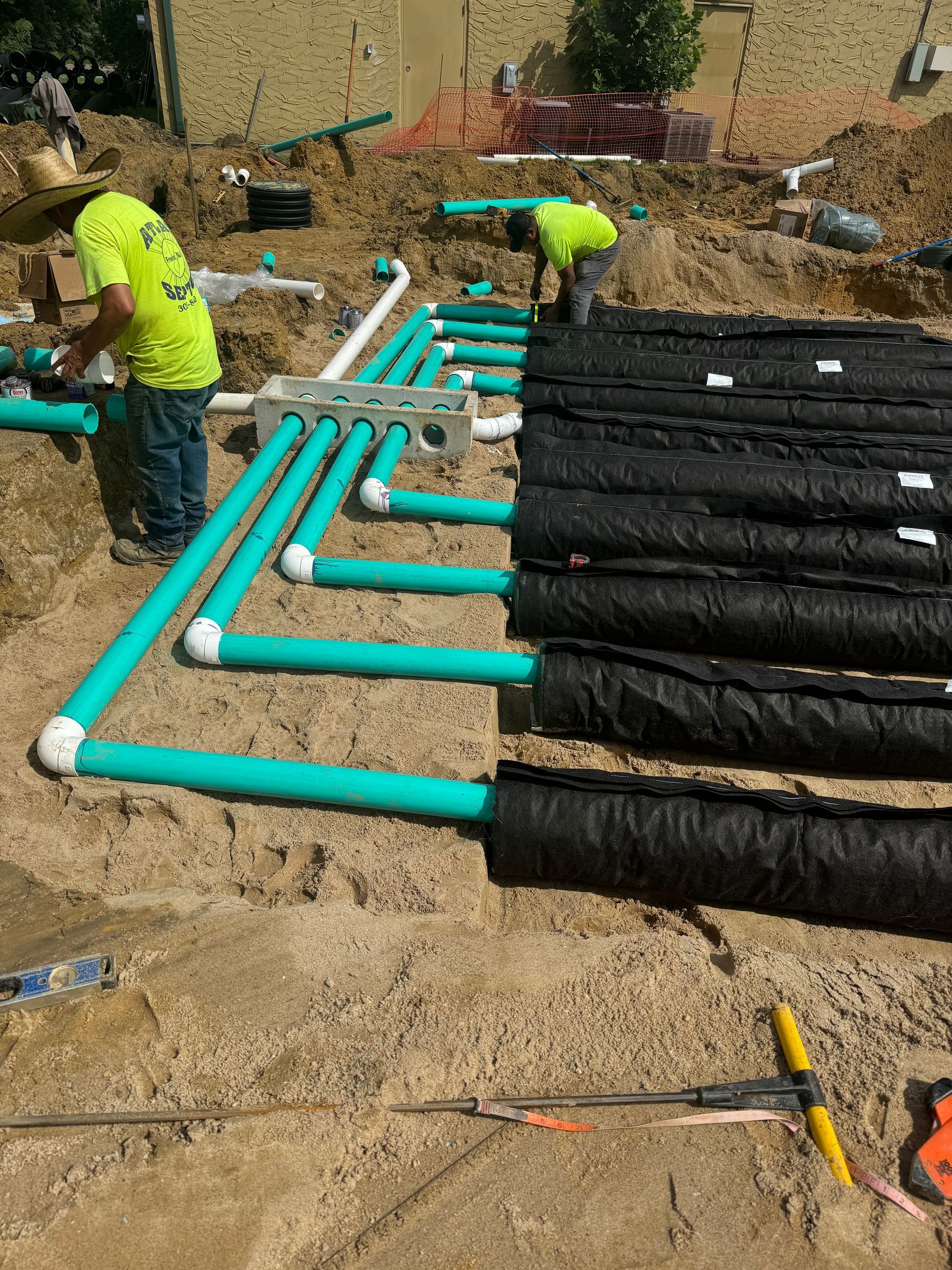 Construction workers installing green pipes connected to black fabric-wrapped objects in a dirt pit.