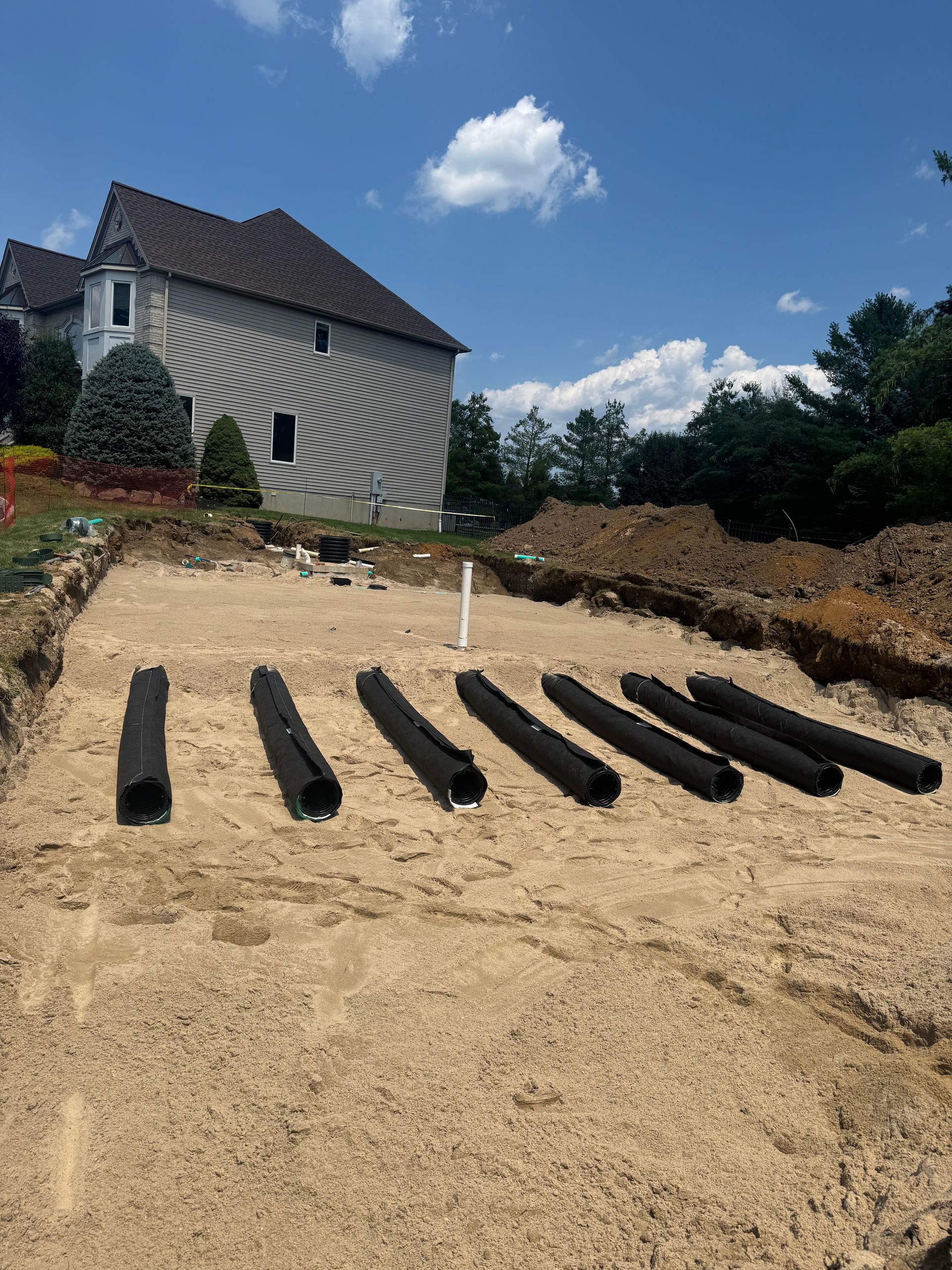 Construction site with buried drainage pipes, house in background, sunny day.