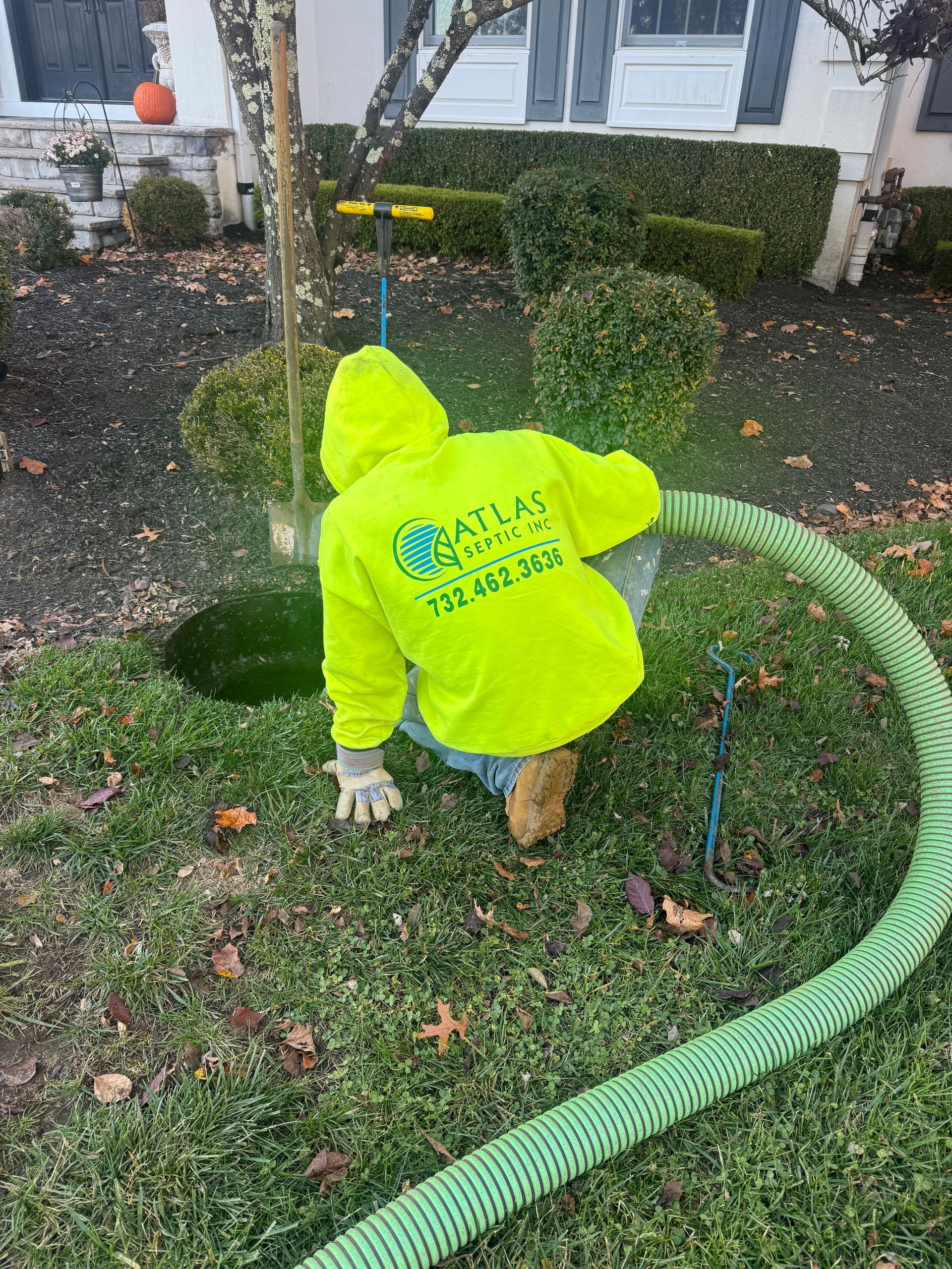 Person in neon yellow jacket servicing a septic tank, with a green hose, in a yard.