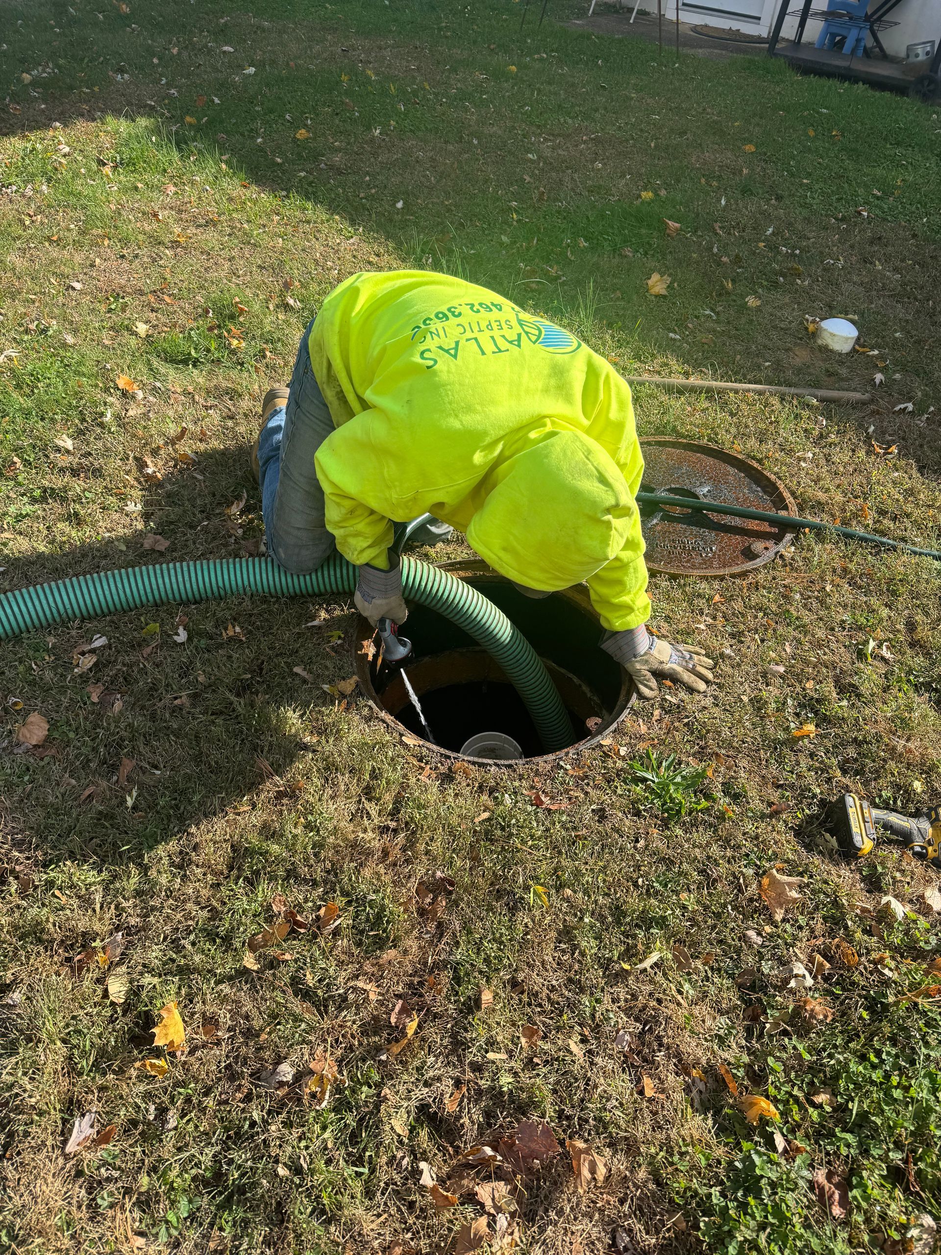 Person in neon yellow hoodie kneeling at an open manhole, with a green hose inserted. Surrounded by grass.