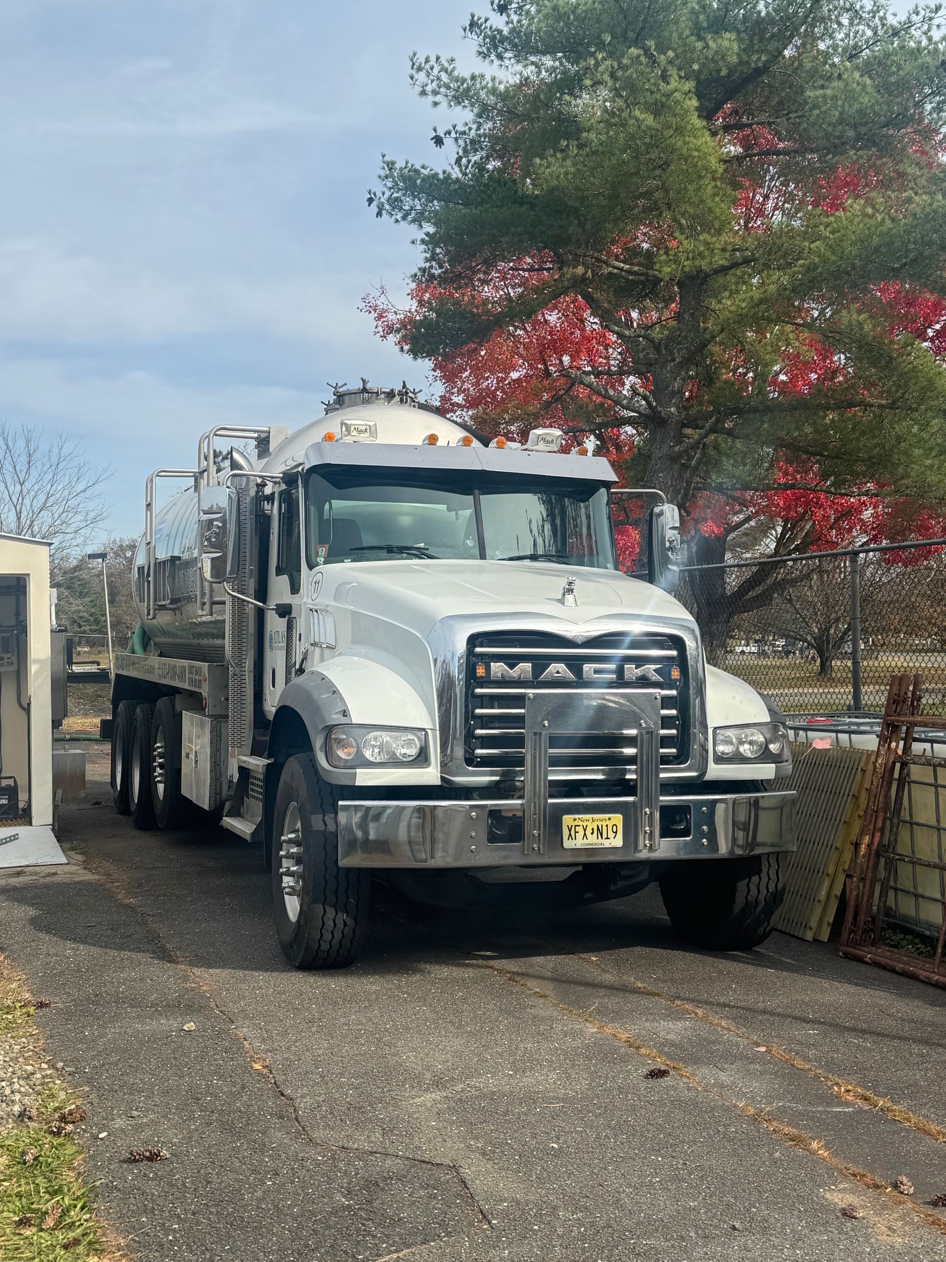Silver Mack truck parked on asphalt driveway, in front of a building and colorful tree.