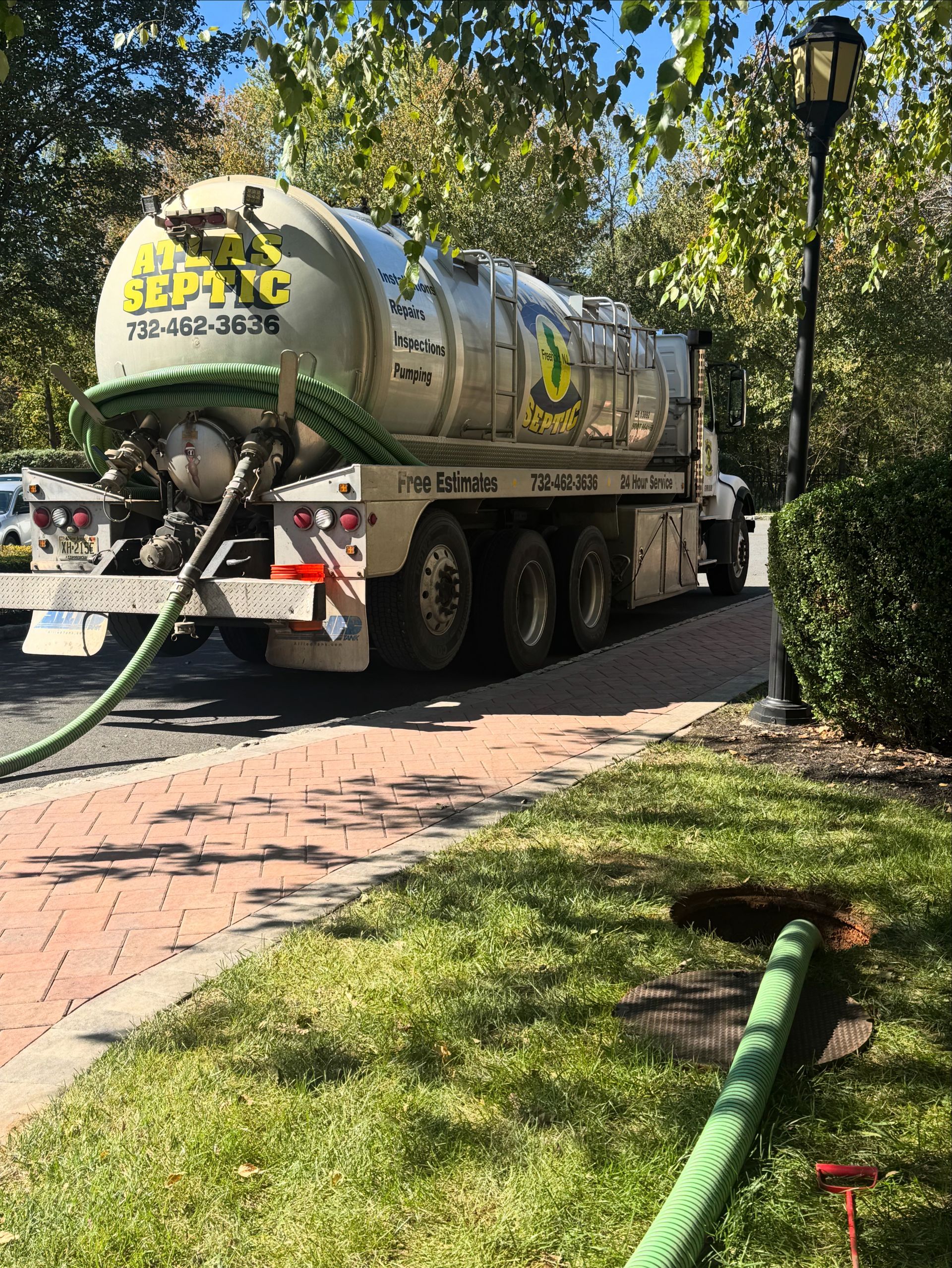 Septic truck parked on a road, connected to a green hose on grass, next to a sidewalk and bushes.
