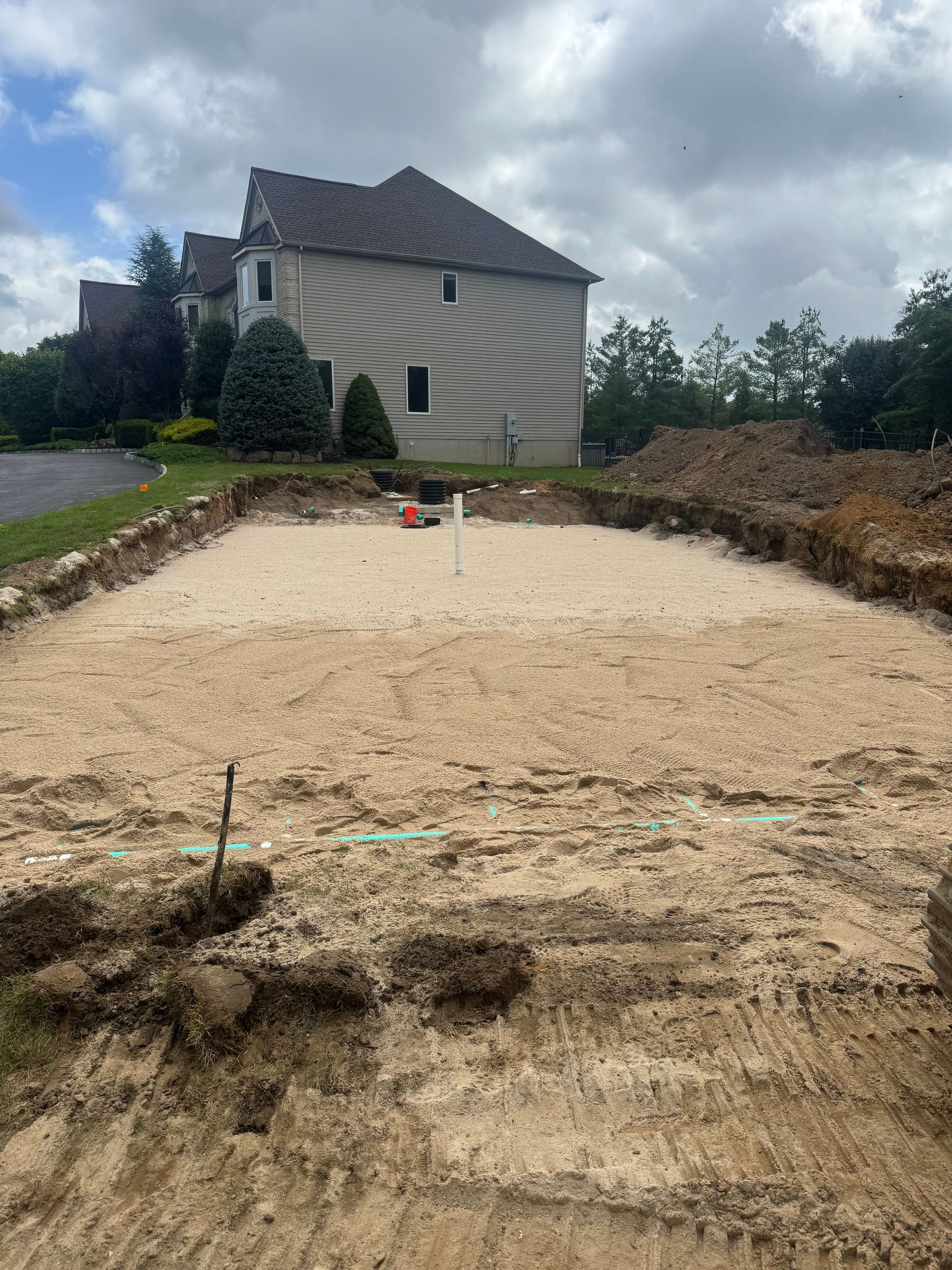 Construction site with rectangular excavation, light-colored soil, and a house in the background.