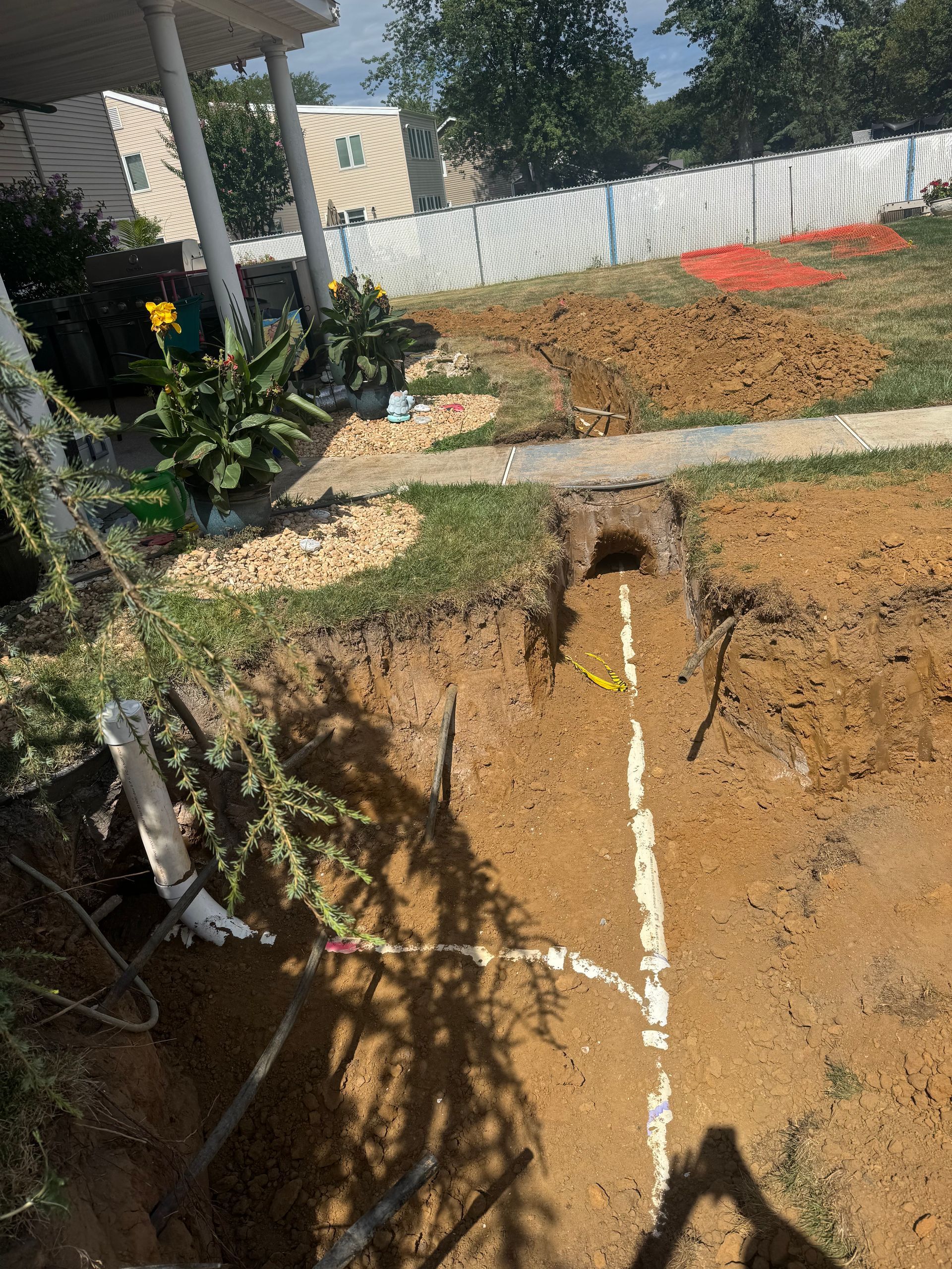 Excavation in progress near a house. White pipe laid in a trench, brown soil, green grass, a fence in the background.