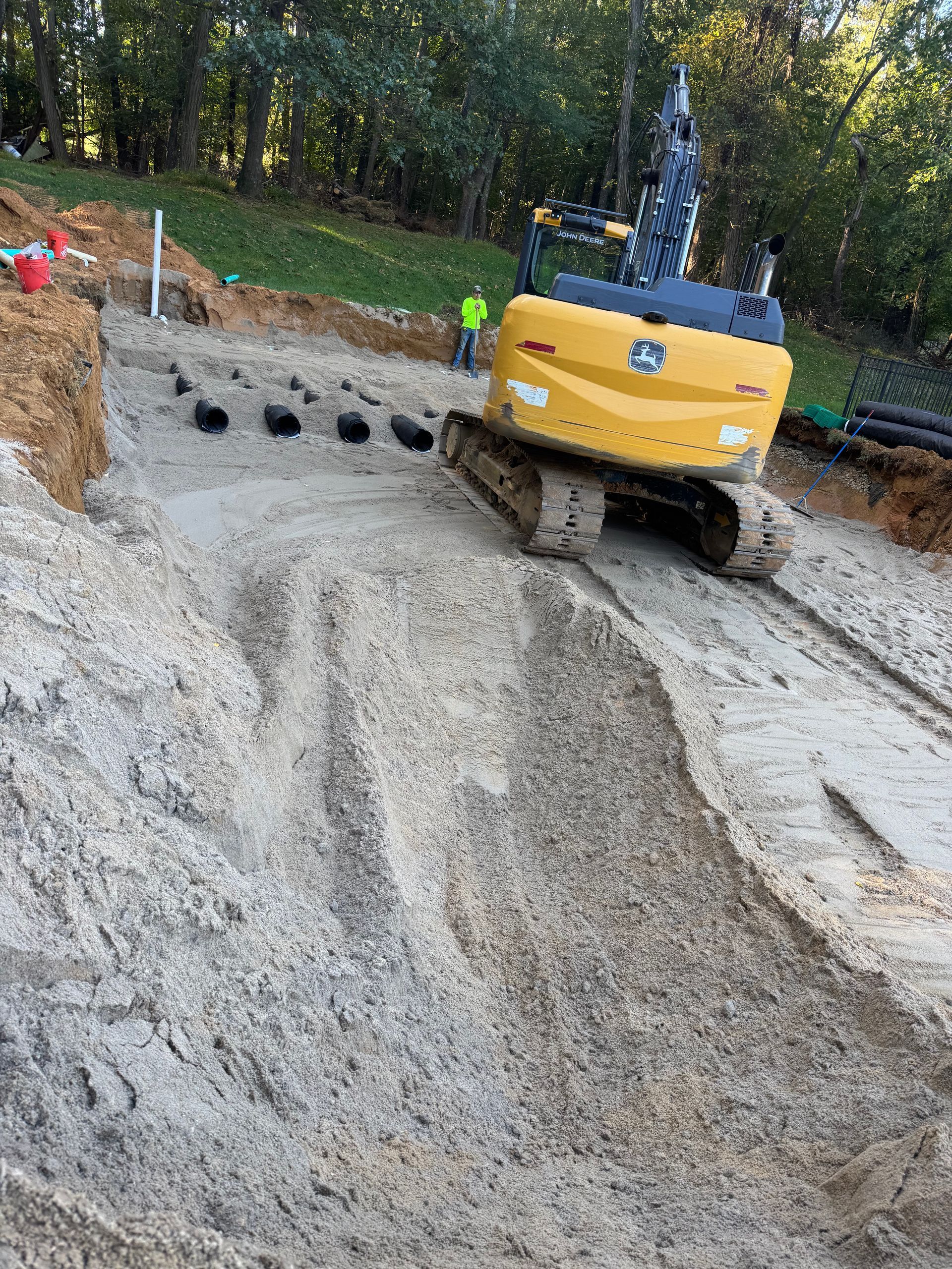 Yellow excavator on a dirt slope near a construction site, with black pipes and a person in a yellow vest.