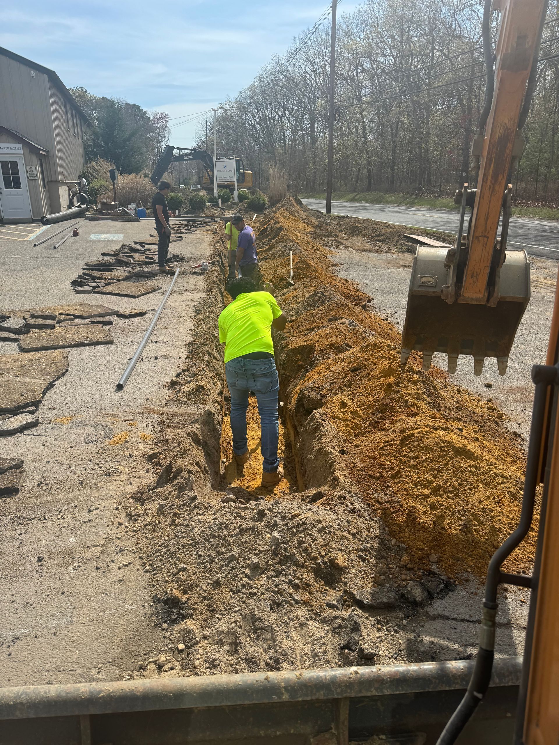 Construction worker in high-vis vest working in a trench, excavator to the right. Roadside setting.