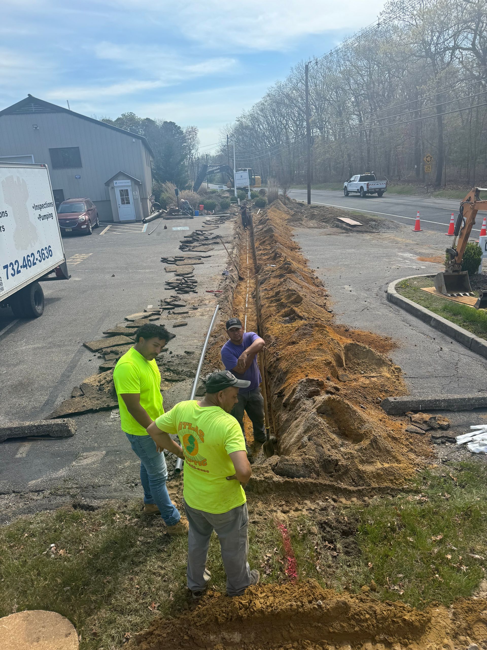 Construction workers in neon shirts standing near an open trench dug in asphalt.