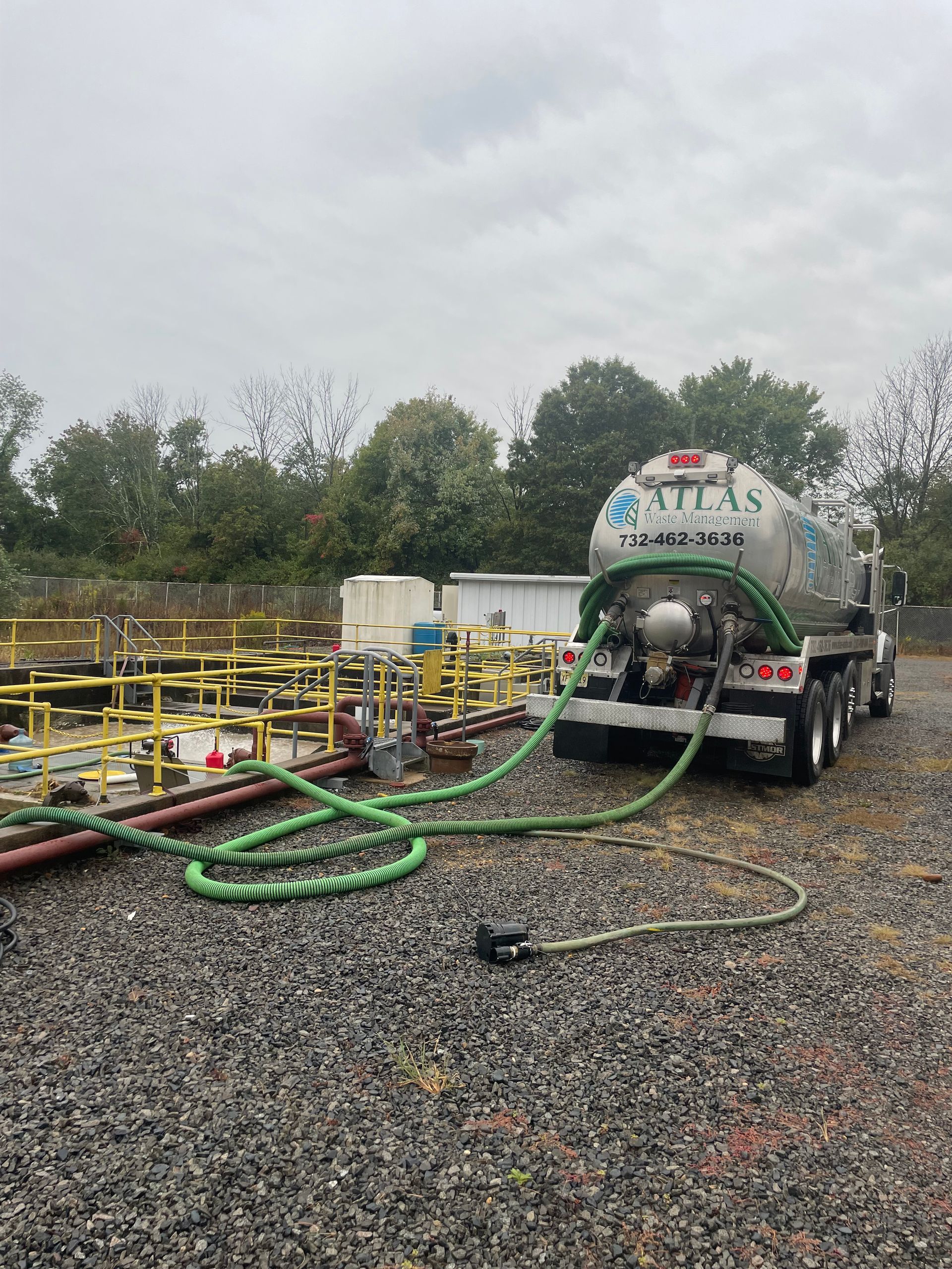 A tanker truck pumps liquid from pipes at a facility. Green hose extends from truck. Overcast sky.