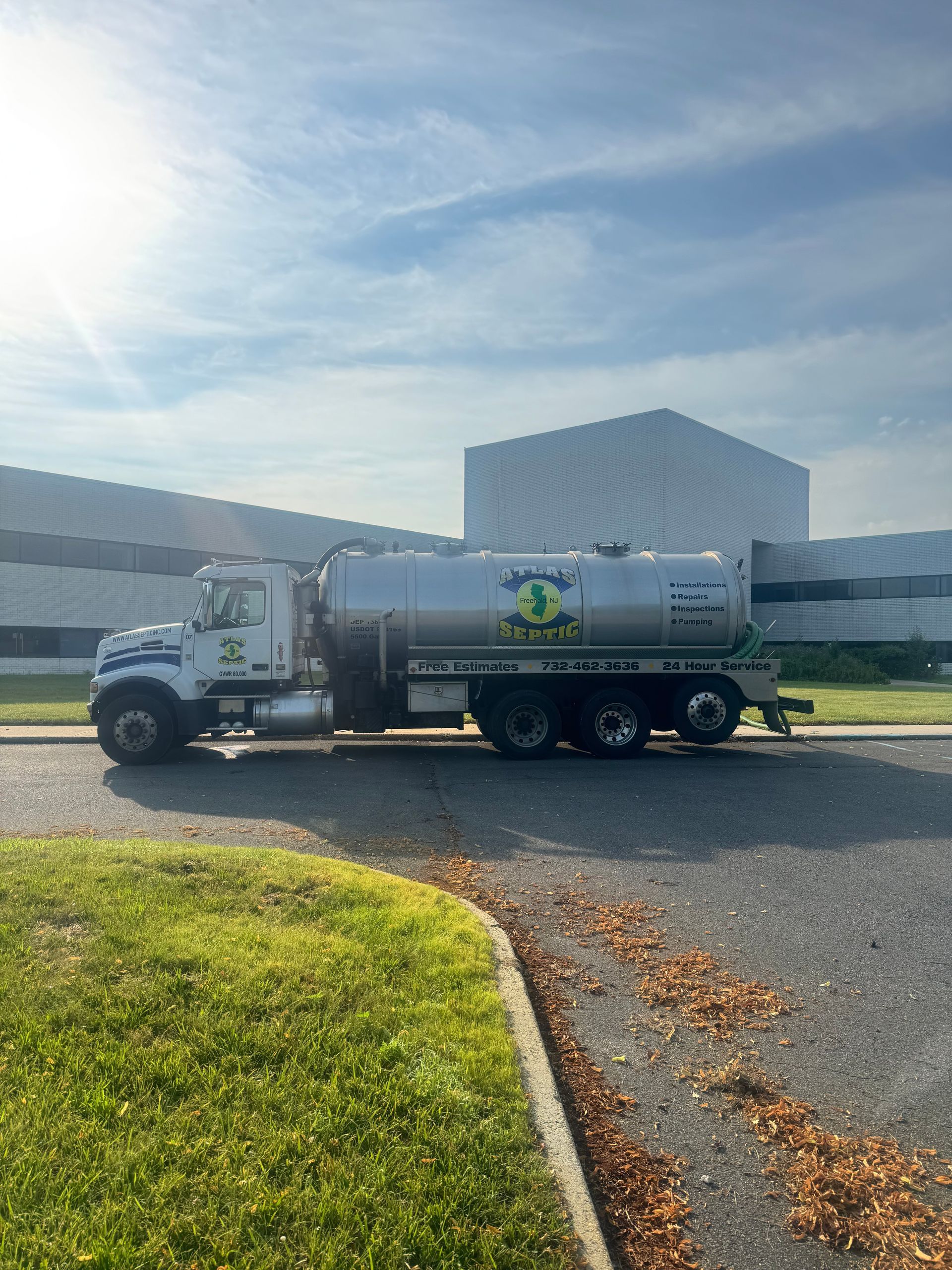A tanker truck parked on asphalt. The stainless steel tank reflects sunlight. Green logo visible.