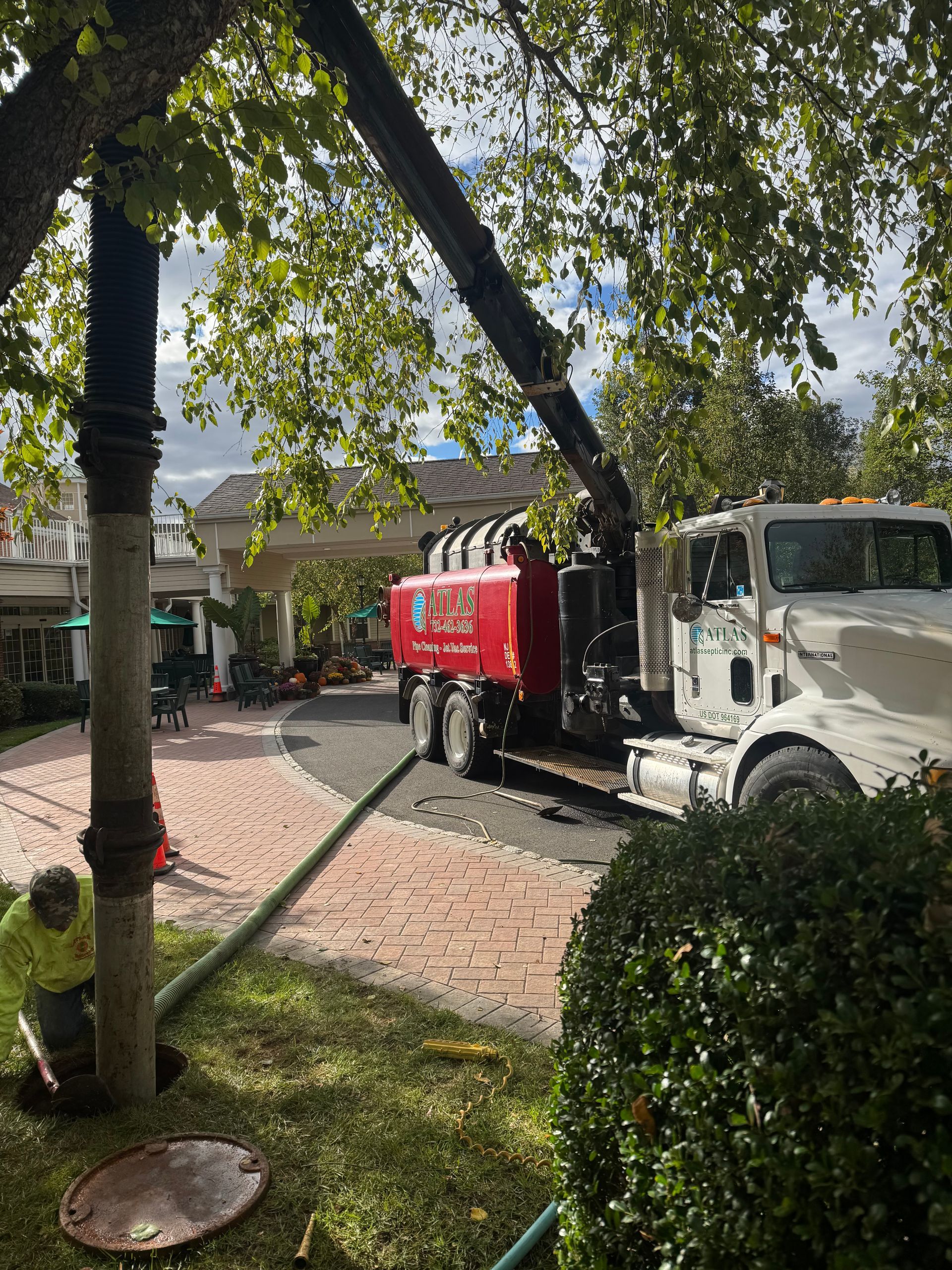 Truck parked near a tree, vacuuming liquid from a manhole on a brick path, with a worker tending to the hose.