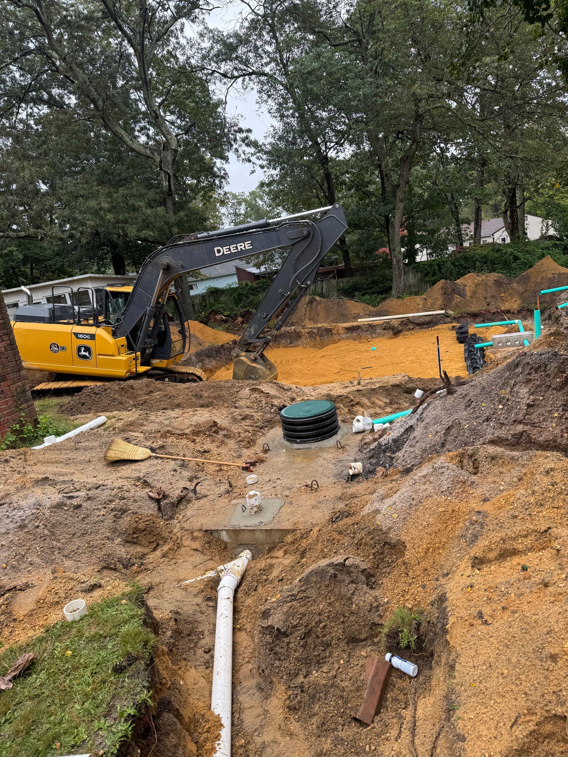 Construction site with excavator digging near a septic tank and pipes. Brown dirt, green grass, and trees.