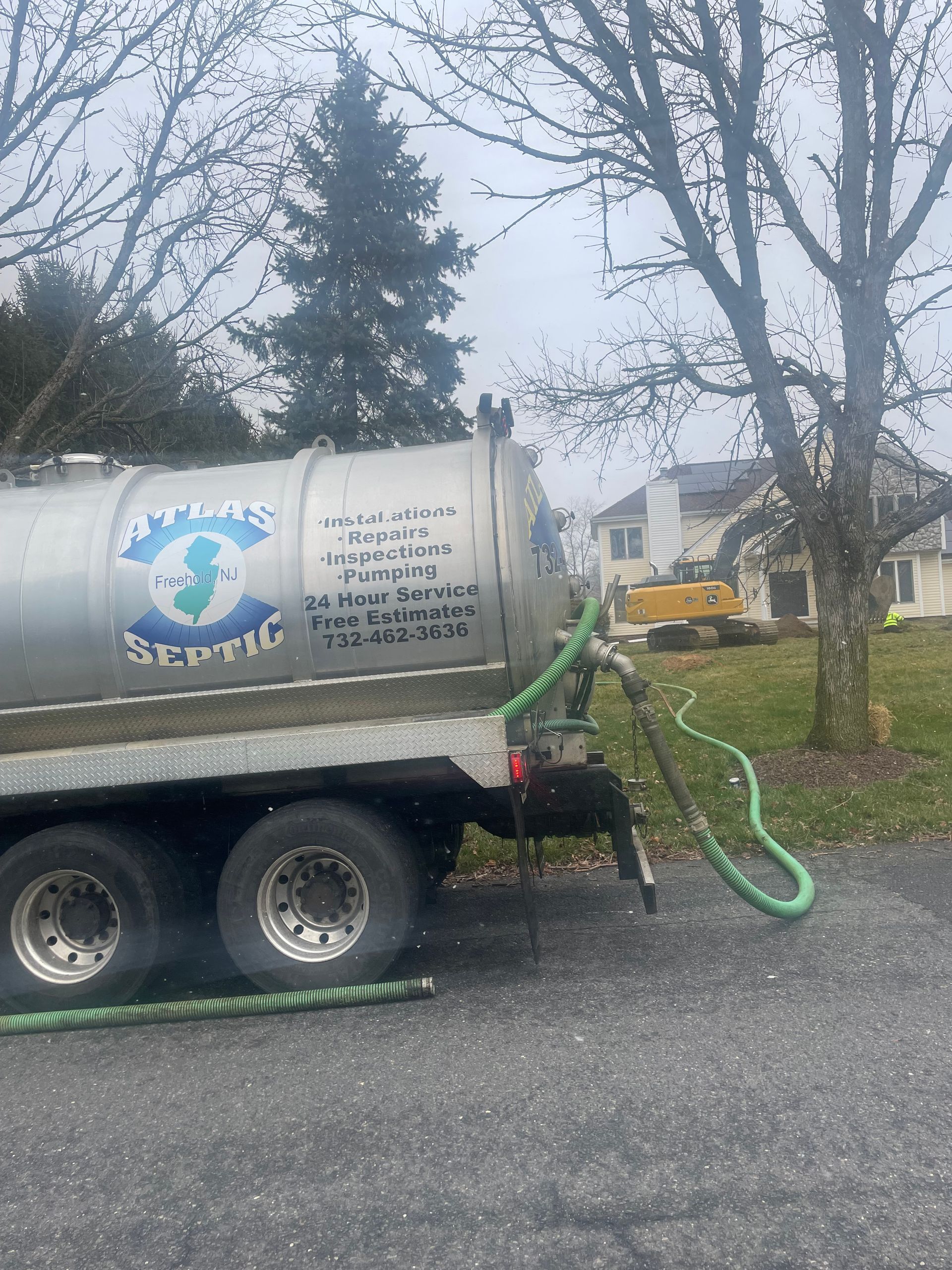 Septic tank truck parked on a road with green hoses. A residential house is visible in the background.