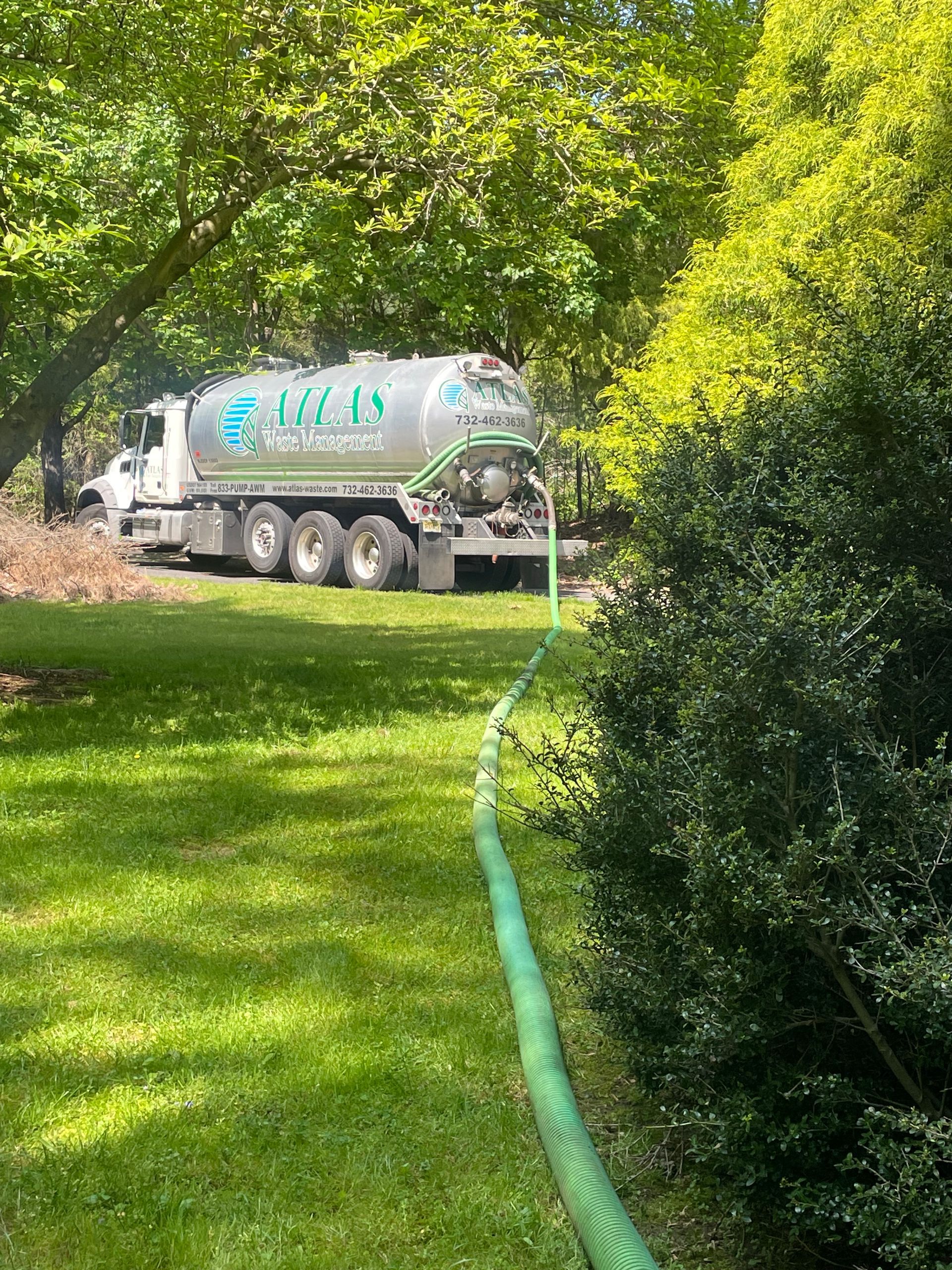 Septic tank service truck on a grassy lawn with a green hose near a bush.