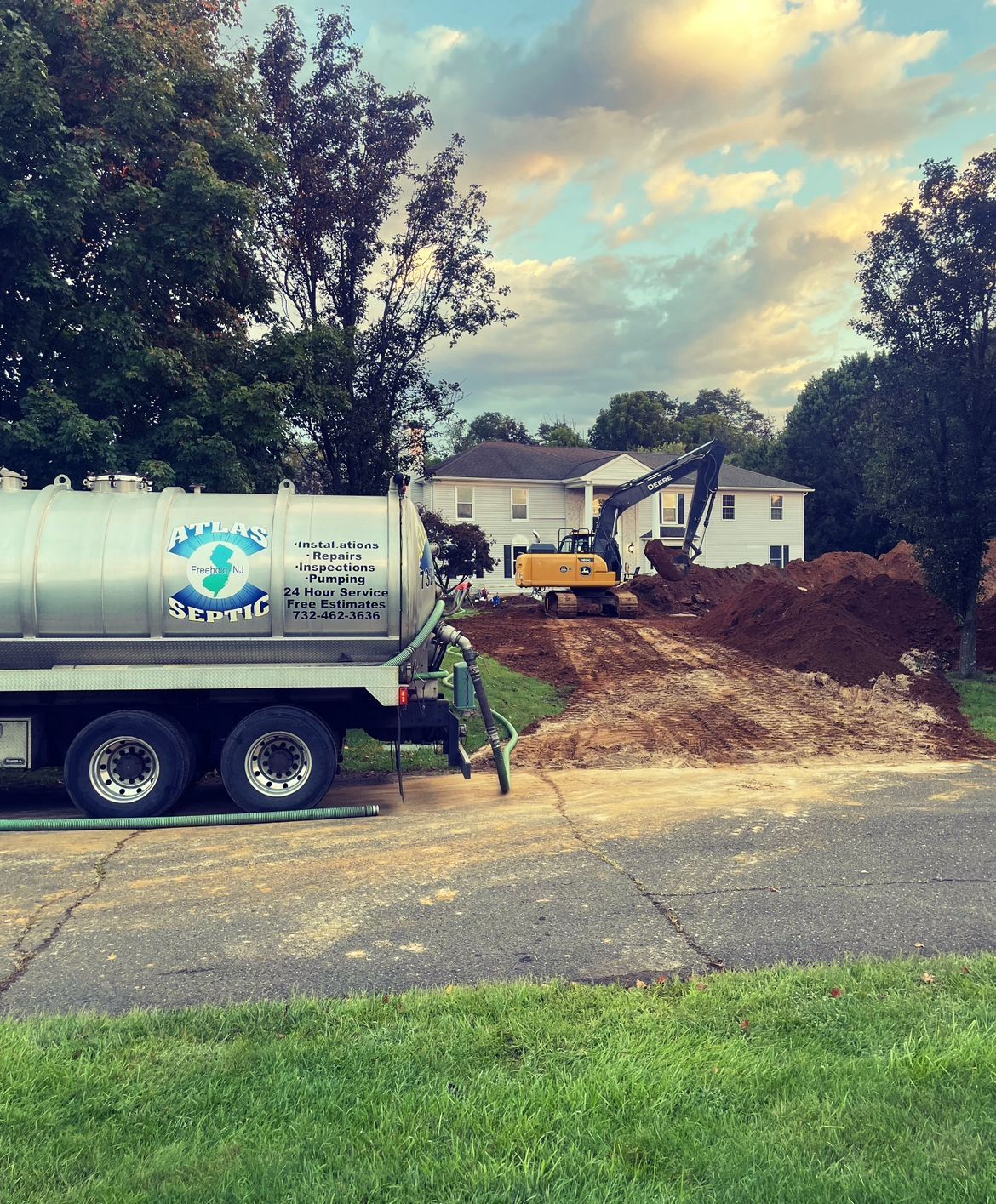 Septic truck and excavator working at a house. Dirt mound in front. Cloudy sky.