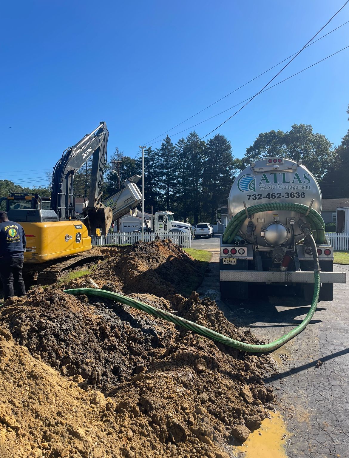 Excavator dumping soil next to a septic pumping truck on a sunny day. Green hose visible.