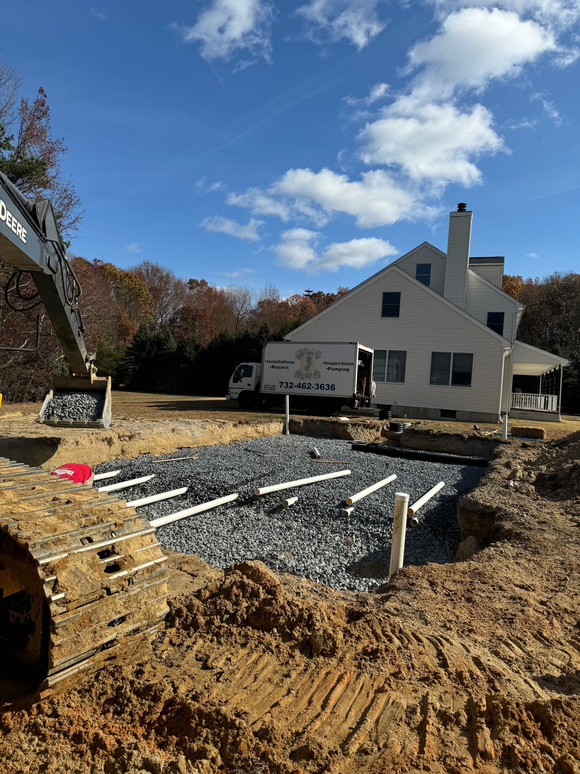 Construction site with a house in the background. Gravel and pipes are in a large pit, excavator arm visible. Blue sky.