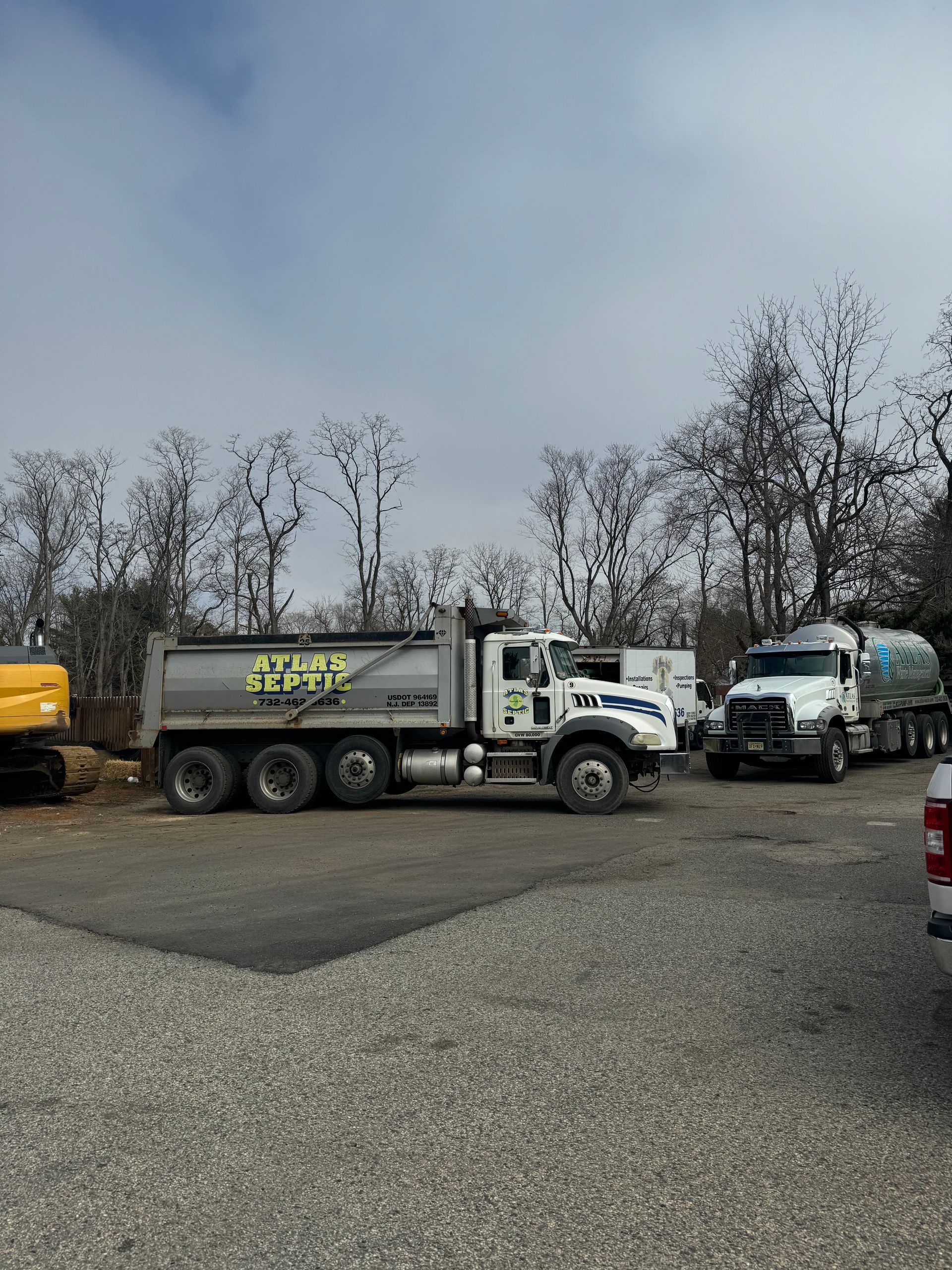 Dump truck and water truck parked on gravel, with trees in the background under a cloudy sky.