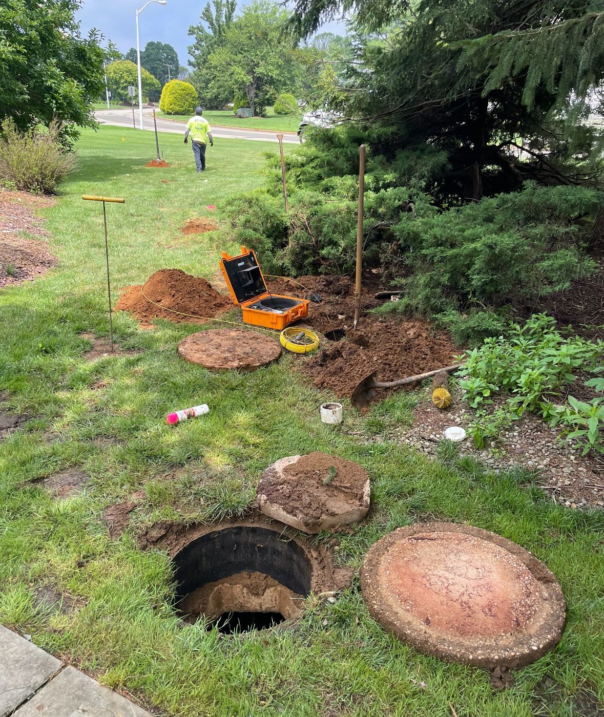 Open utility hole in grass with nearby dirt piles, tools, and a worker in the background.