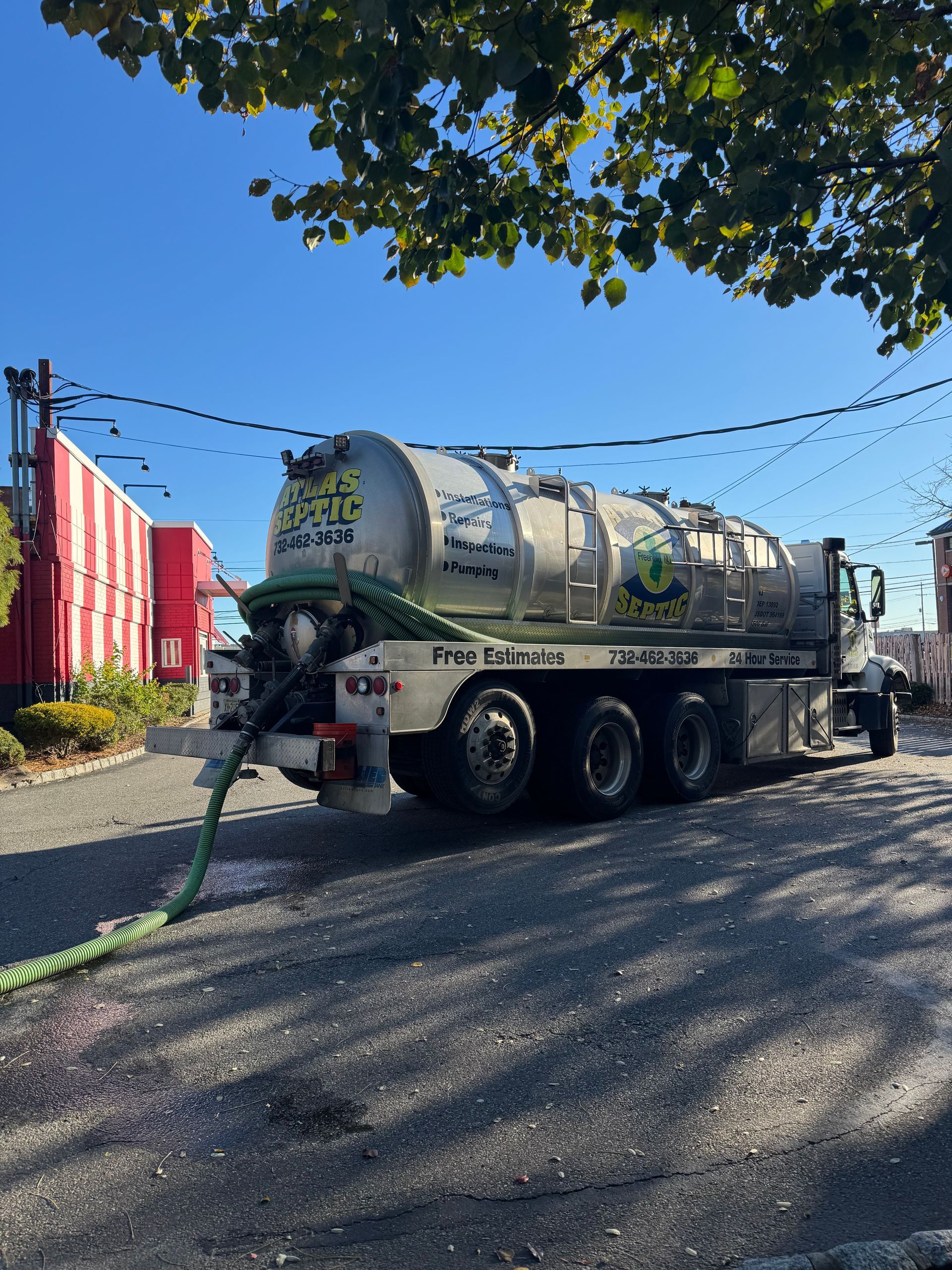 Septic service truck with green hose in use. Gray tank, parked on asphalt, sunny day.