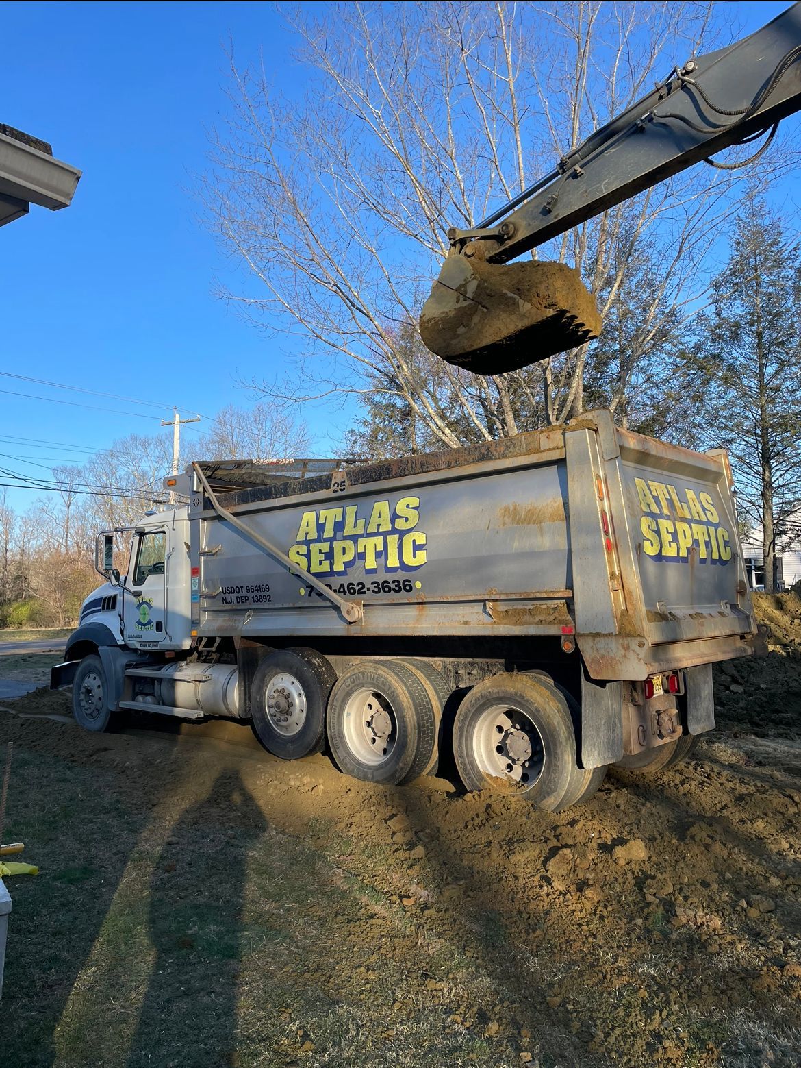 Excavator dumping soil into an Atlas Septic dump truck on a sunny day.