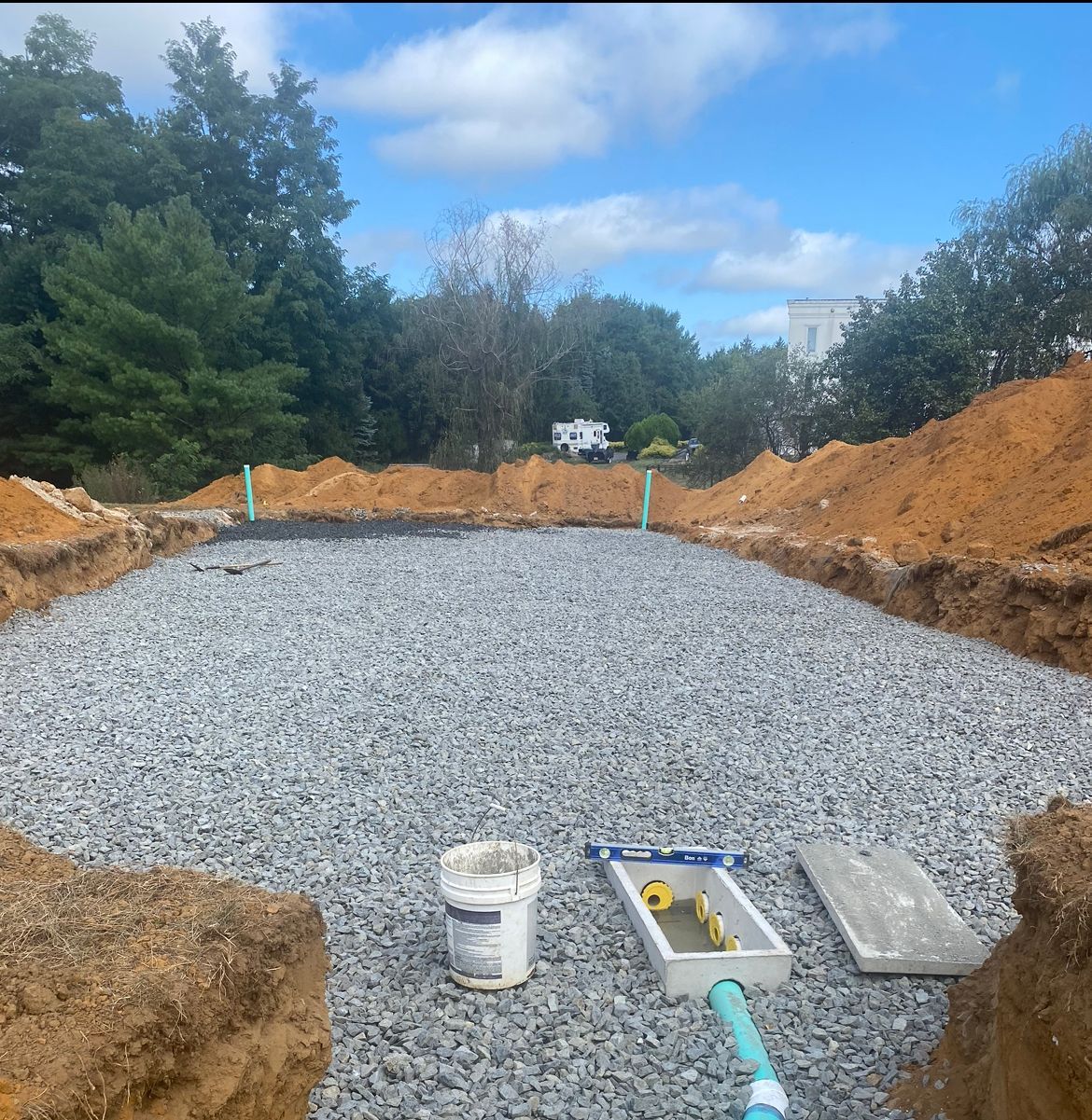 Gravel-filled rectangular pit with PVC pipes, tools, and a white bucket. Excavation area with trees and blue sky.