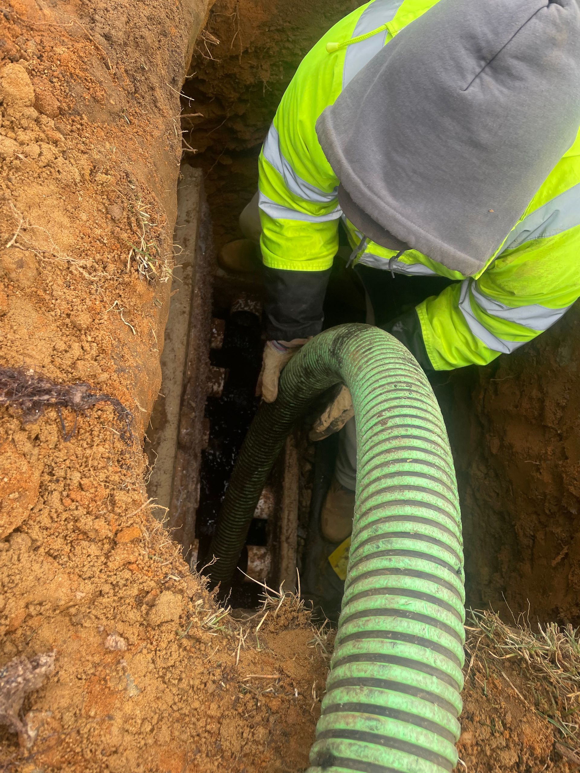 Person in a high-visibility jacket pumps a green hose into a dark opening in a muddy excavation.
