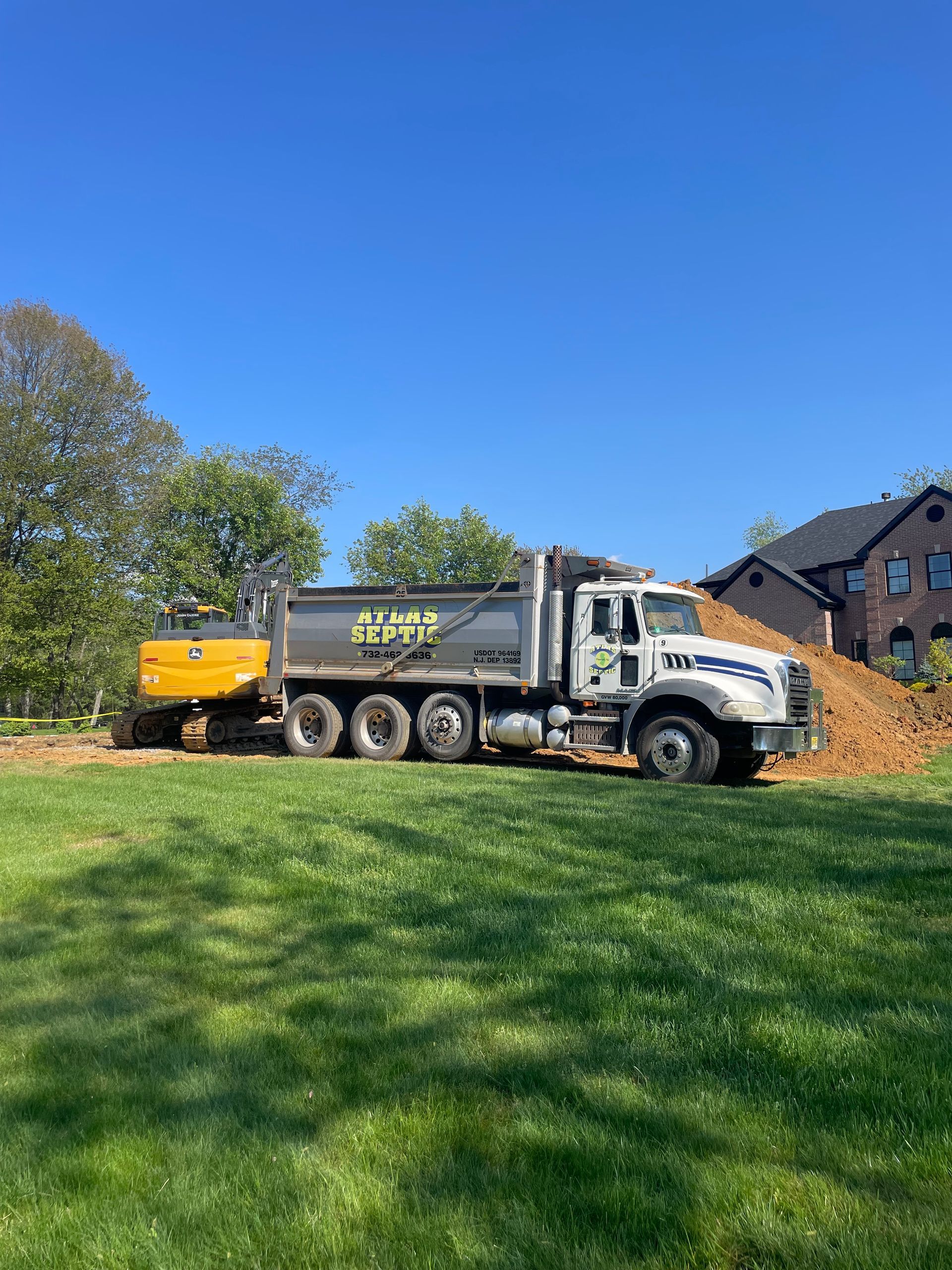 Excavator loading dirt into dump truck on a grassy lawn with a house in the background. Bright blue sky.