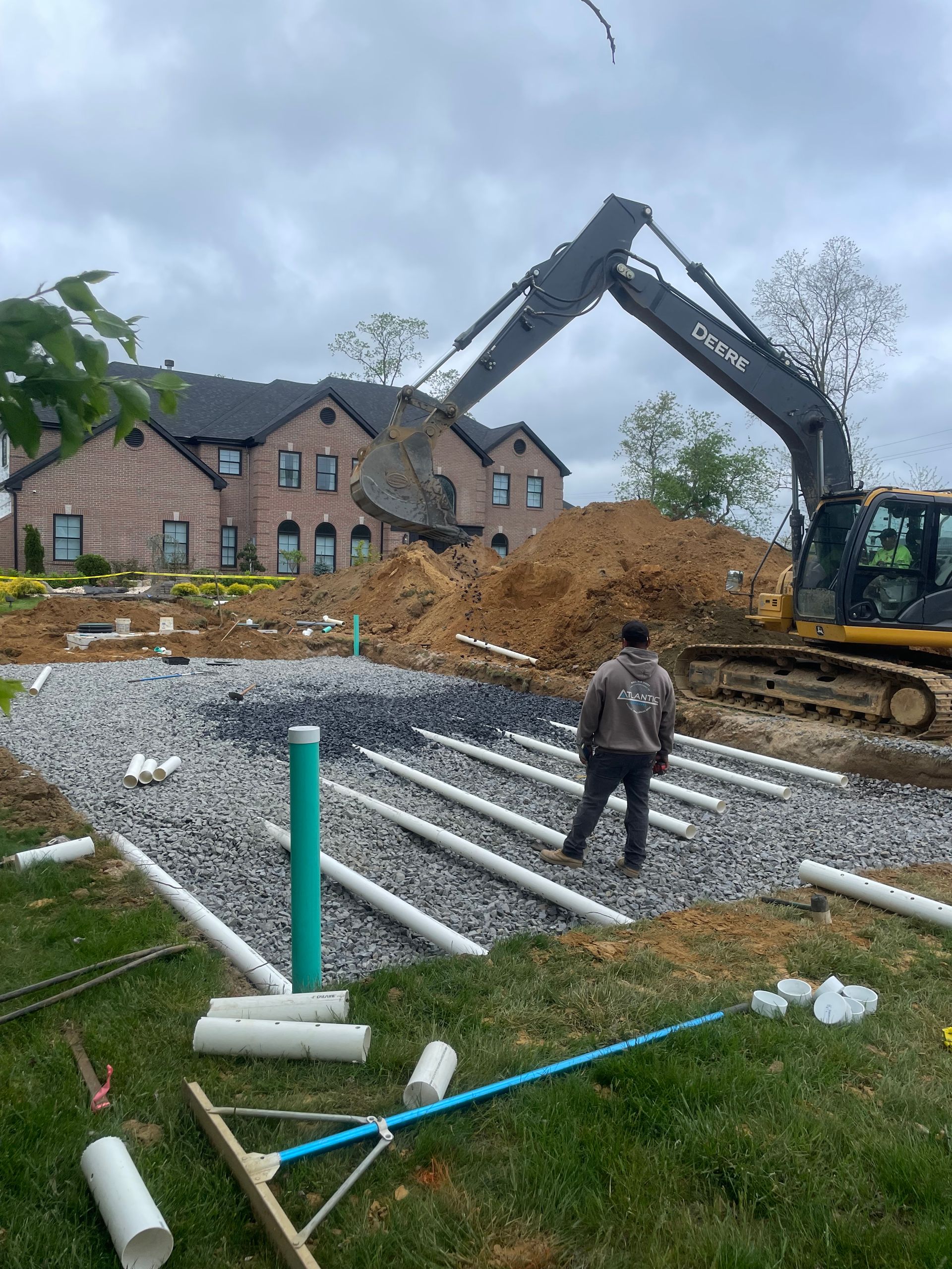 Construction site with a backhoe, gravel bed, pipes, and worker; house in background.