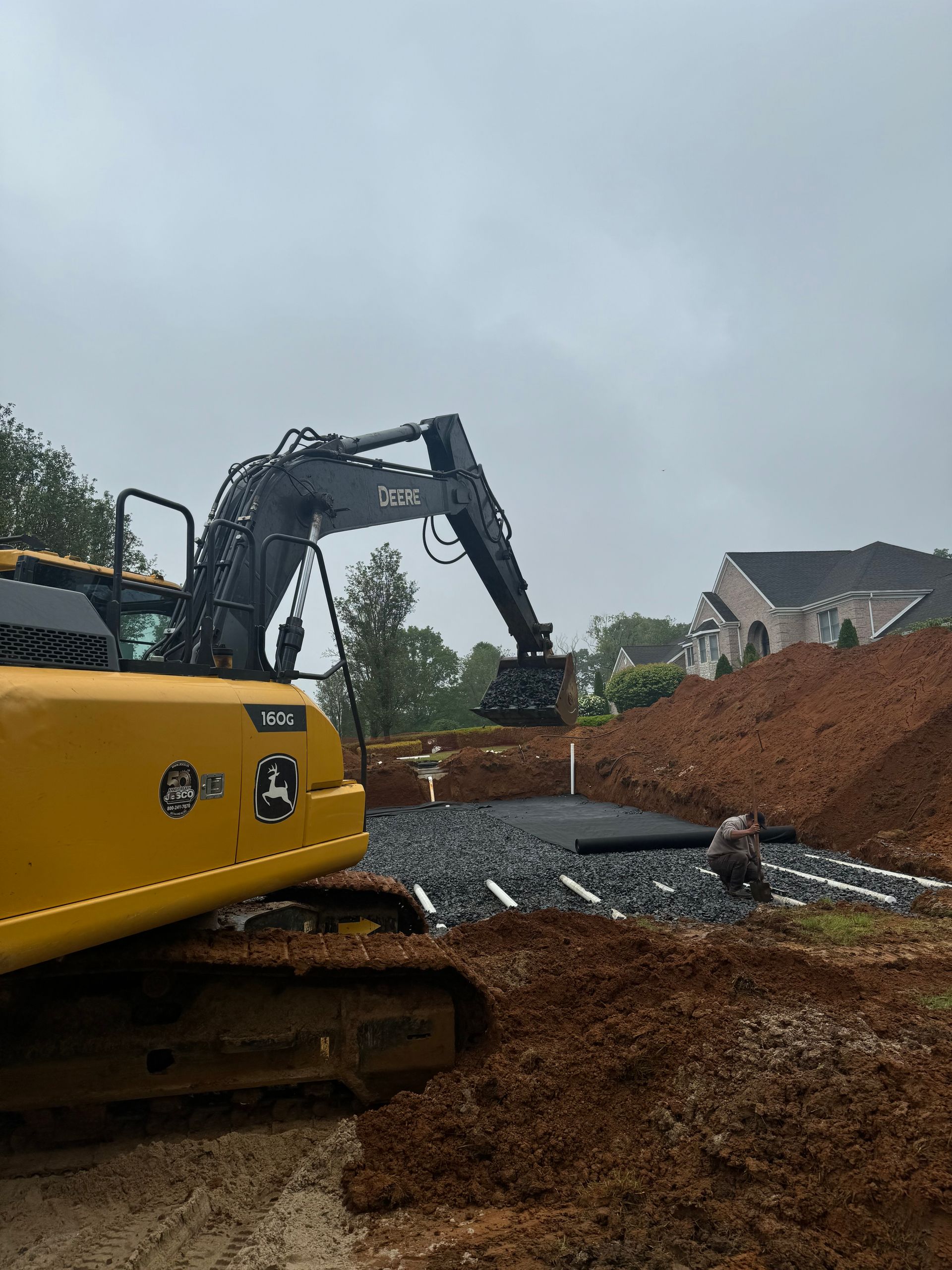 Yellow excavator digging in a construction site with exposed pipes and a residential building in the background.