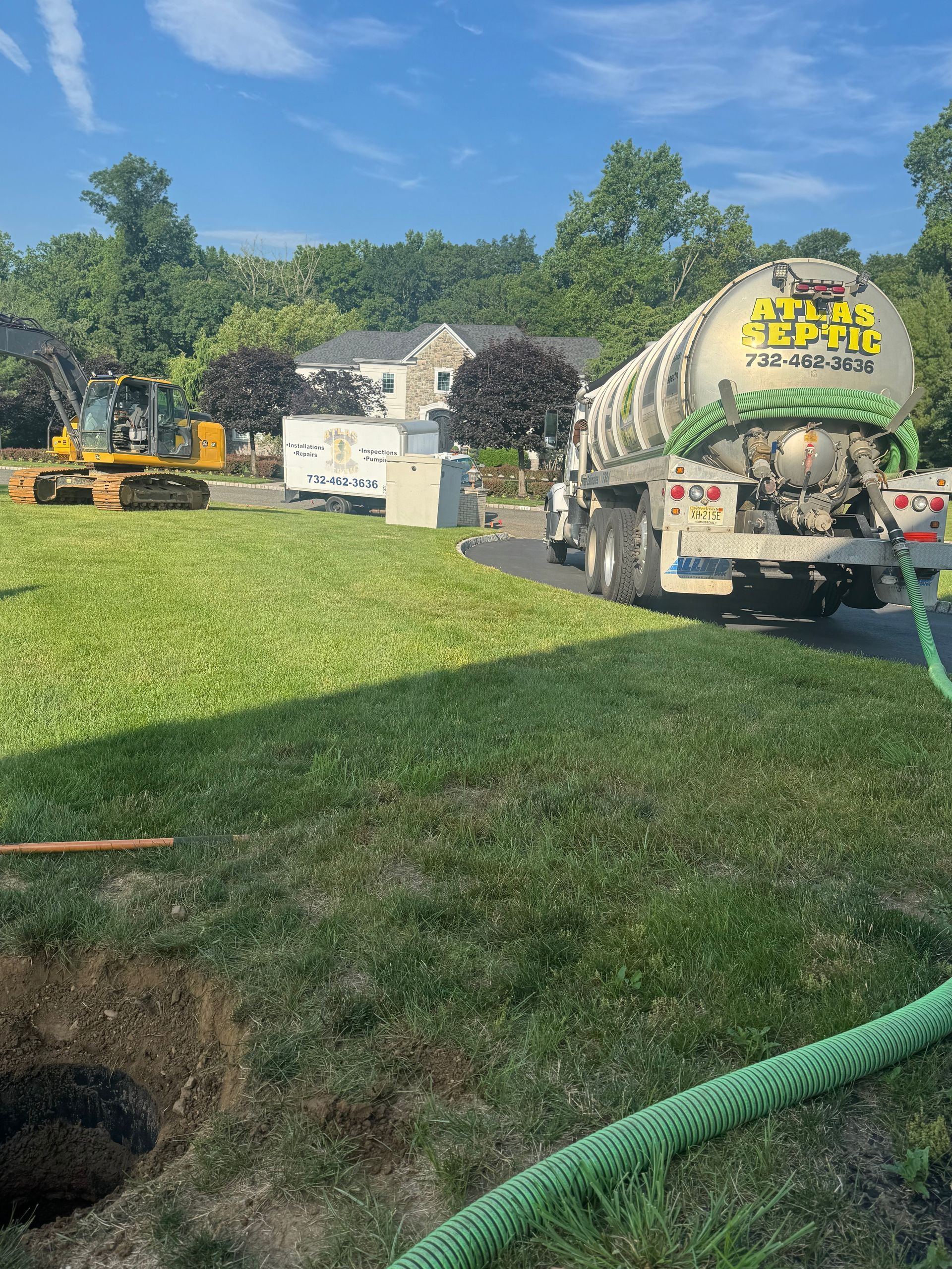 Septic tank truck and excavator on a grassy lawn; a hole in the foreground.