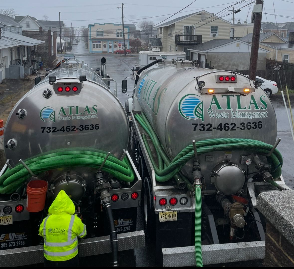 Two Atlas septic trucks parked on a street; a worker in yellow rain gear near one truck.