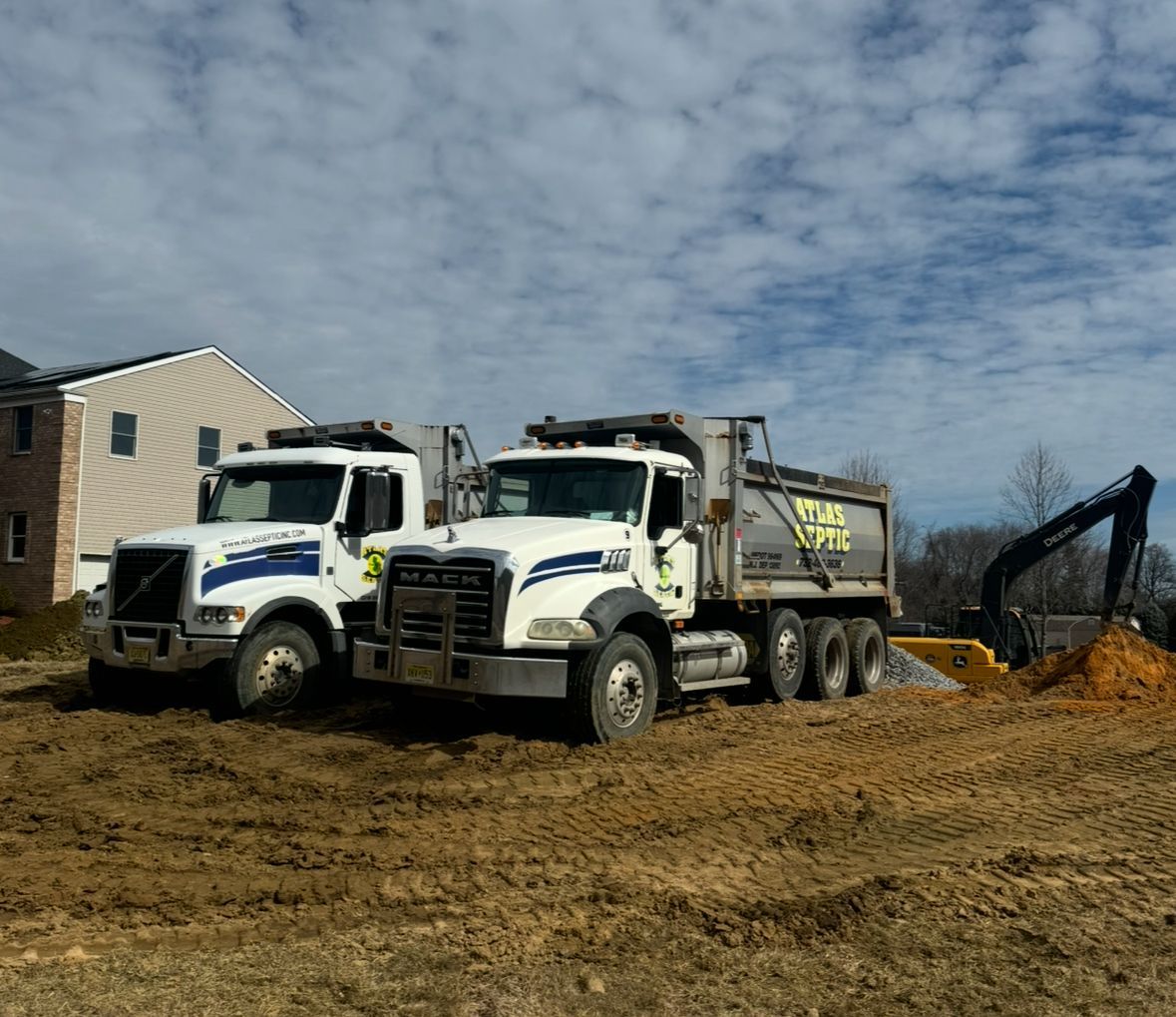 Two white dump trucks parked on a dirt lot with an excavator in the background. Cloudy sky.