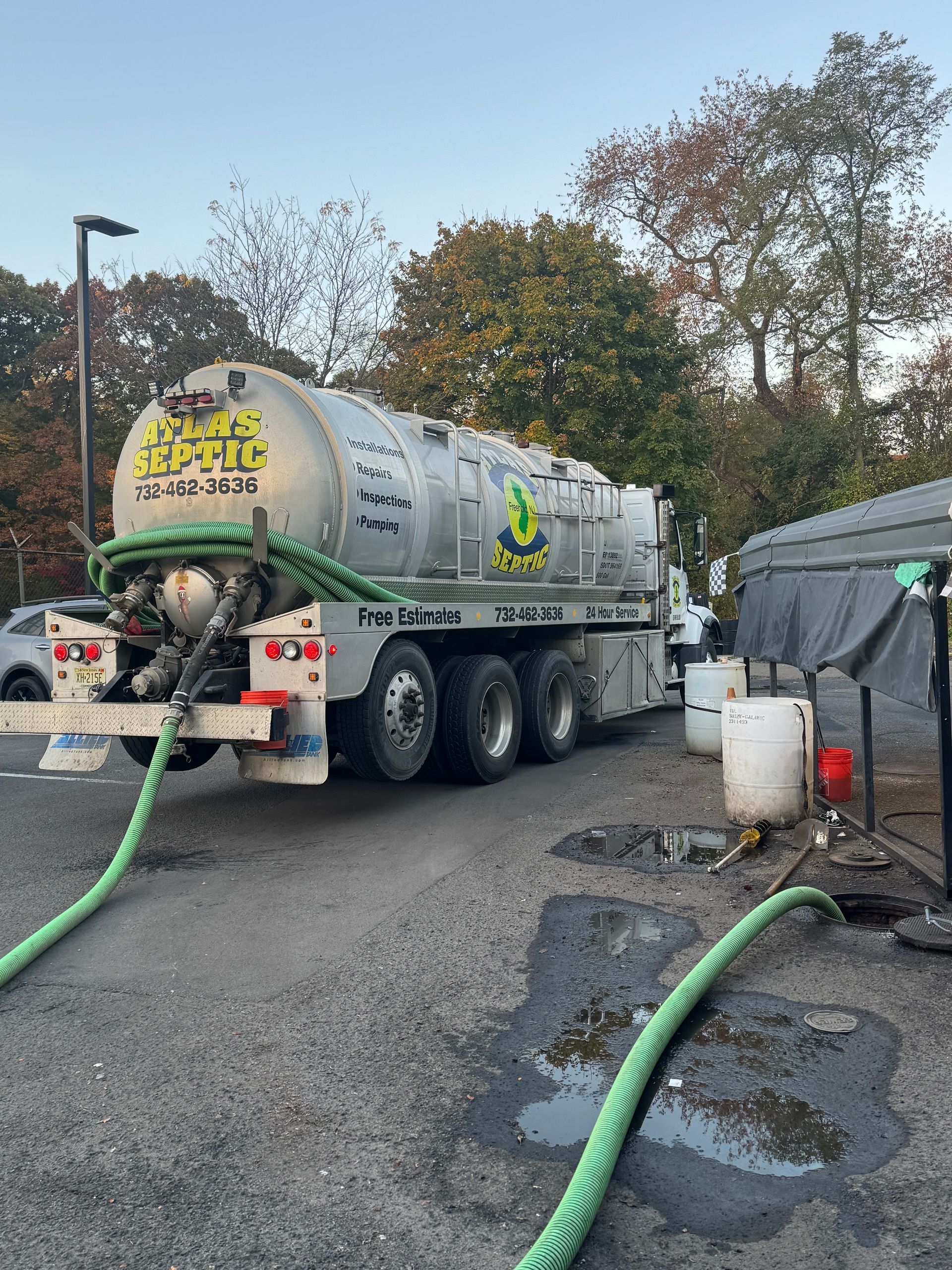 A large silver septic tank truck parked in a lot, connected to hoses, with trees in the background.