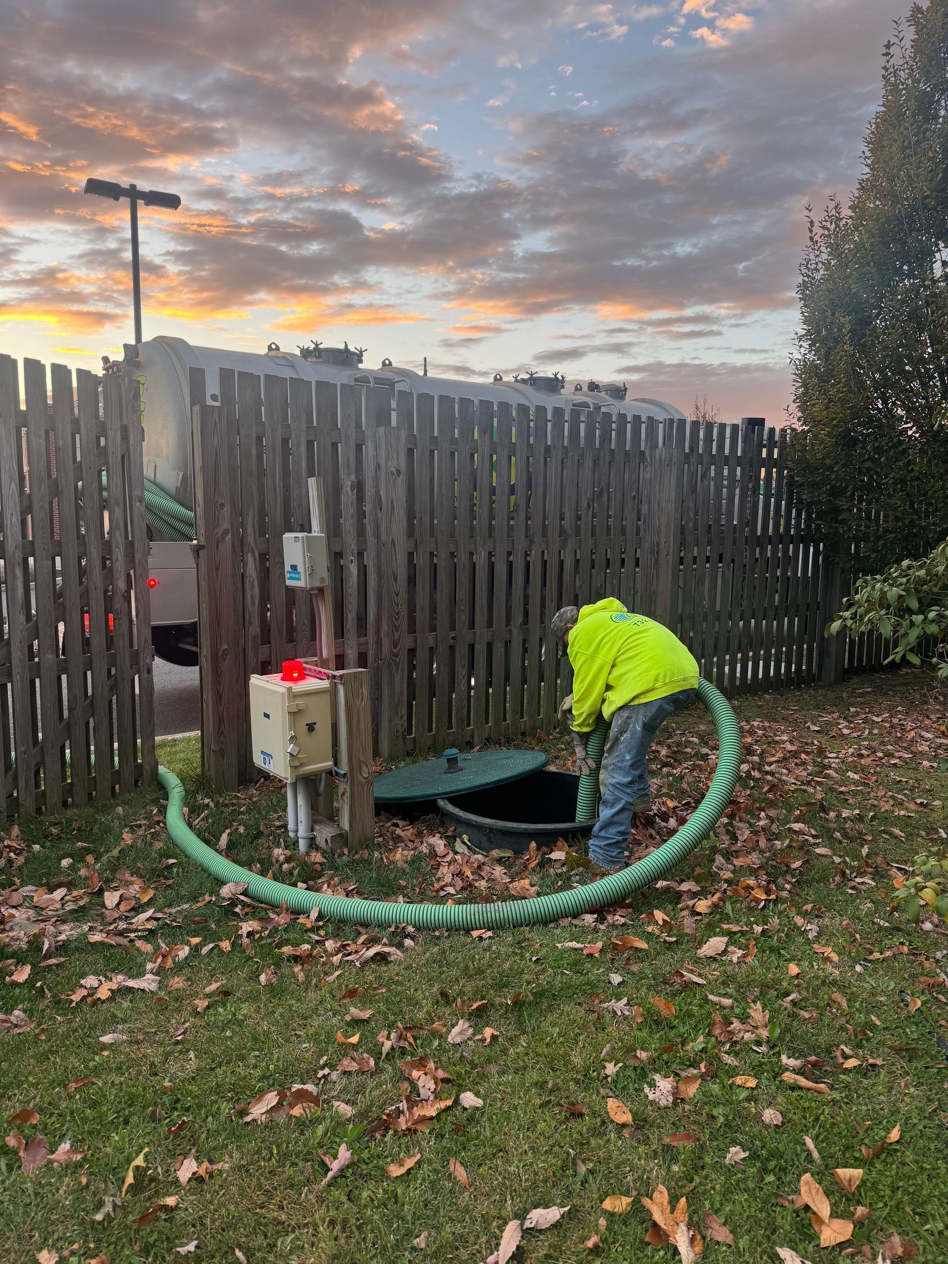Man in a yellow safety vest pumps a septic tank behind a wooden fence. Cloudy sunset in background.