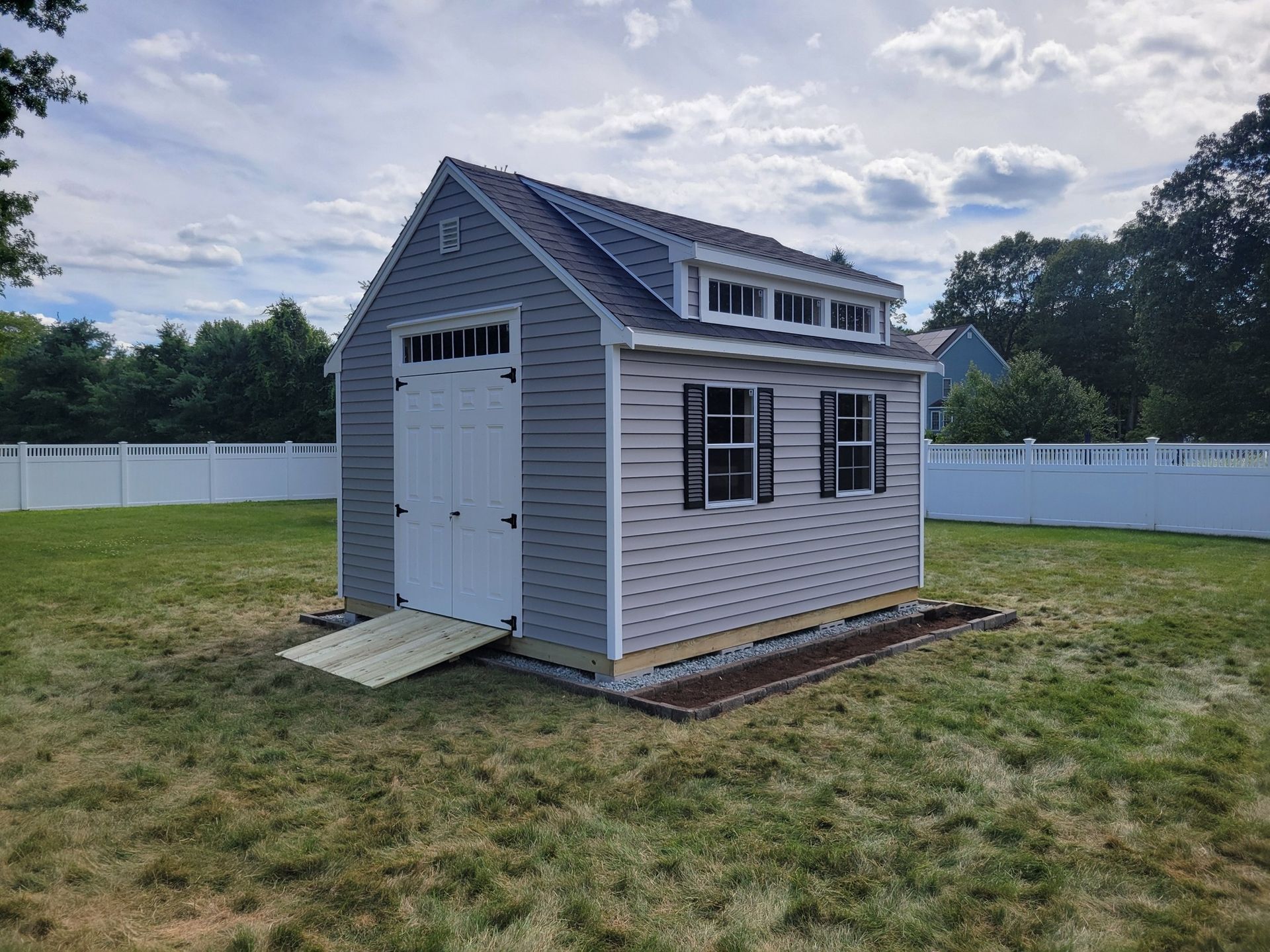 Gray shed-style garage with white doors and shutters on a grassy lawn under a cloudy sky