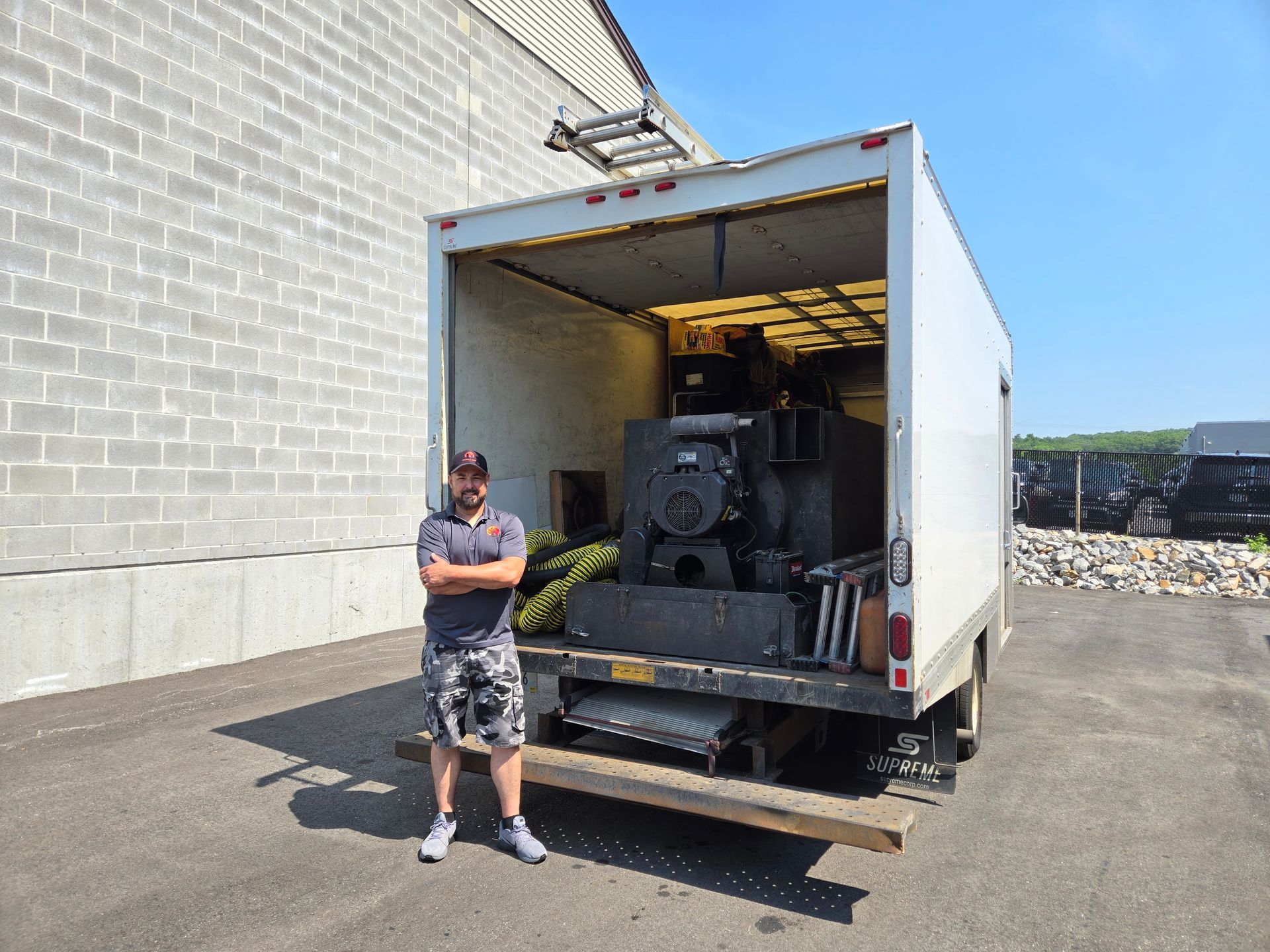 A man stands by a truck containing large equipment; he has crossed arms. Outdoors, sunny day.