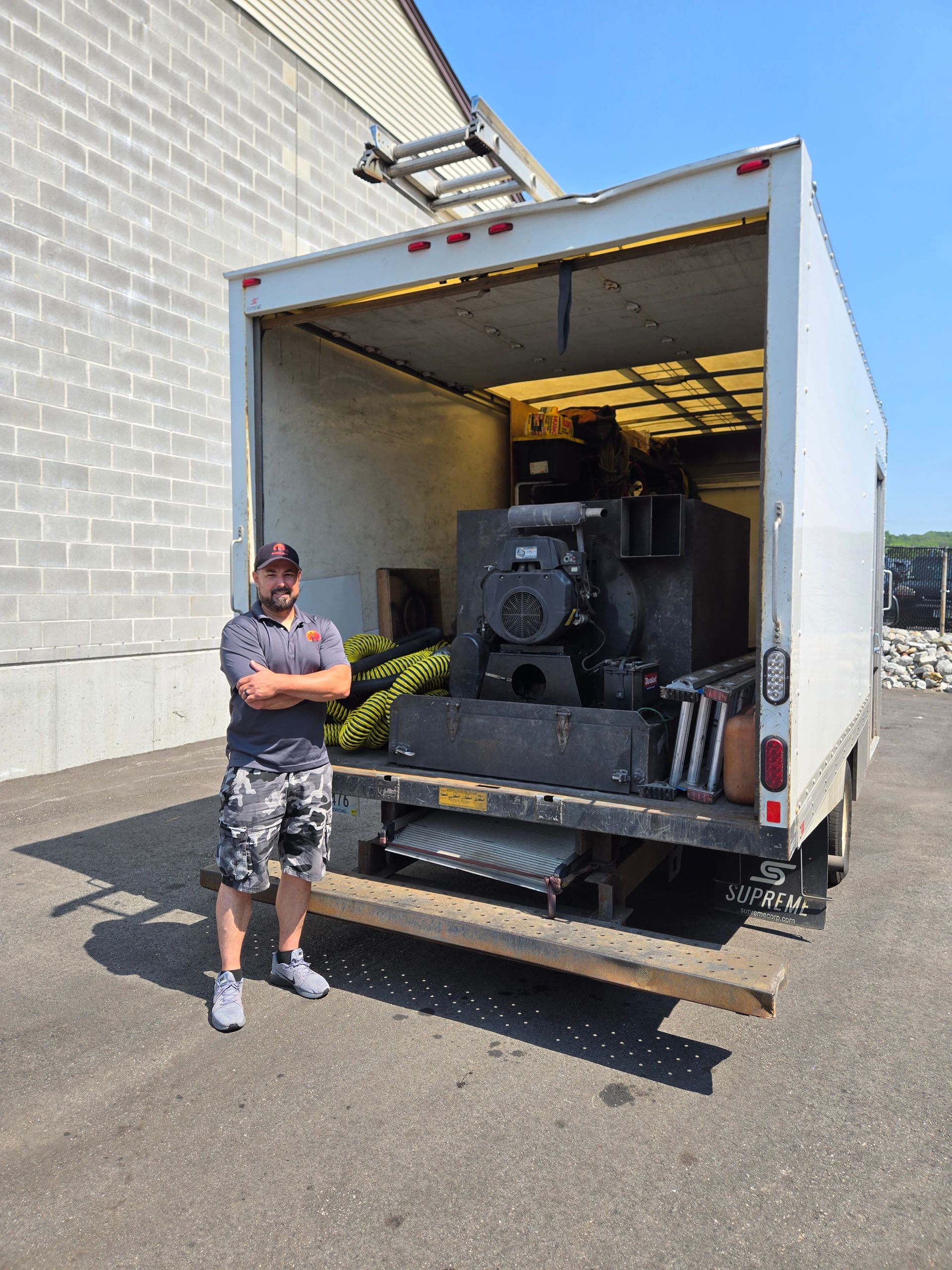 A man stands in front of a truck loaded with equipment, on a sunny day in a loading area.