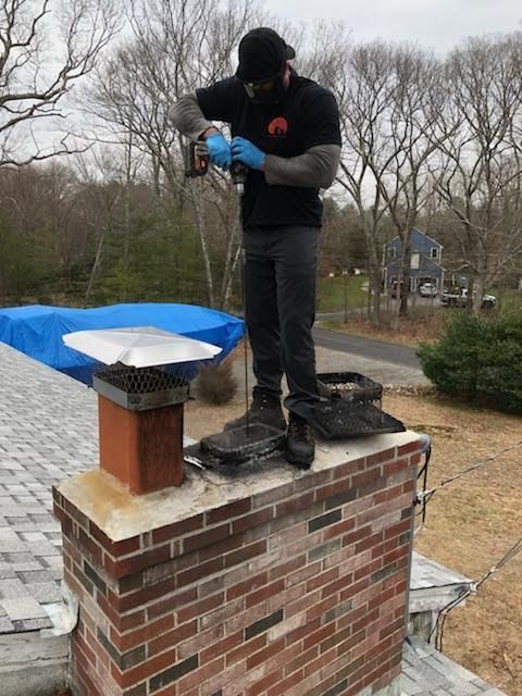 Man on a brick chimney, using a power tool. Gray sky, trees, and a blue tarp nearby.