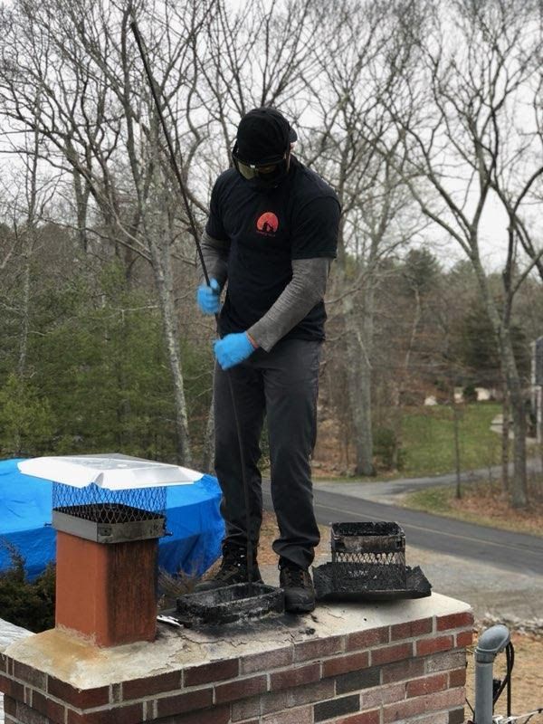 Chimney sweep on a roof wearing blue gloves, cleaning a brick chimney with a long brush.