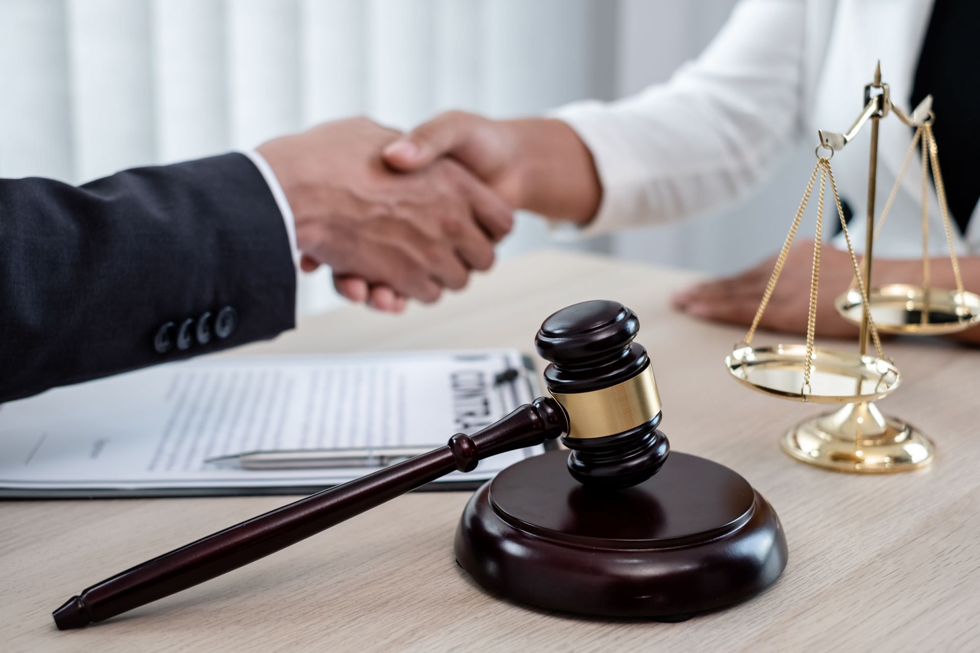 Two people shaking hands over a contract with a wooden gavel and a gold scale of justice on a desk.