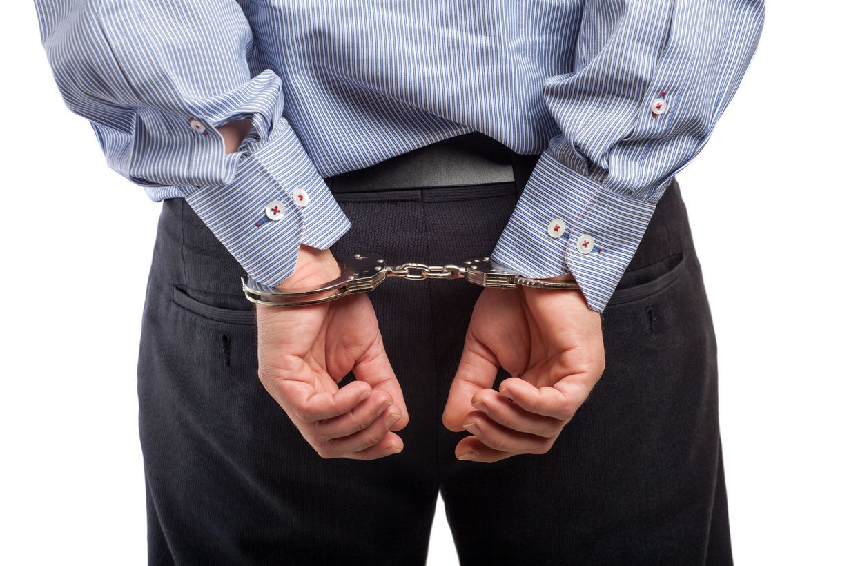 A person in a blue pinstriped shirt and dark trousers stands with their hands cuffed behind their back.