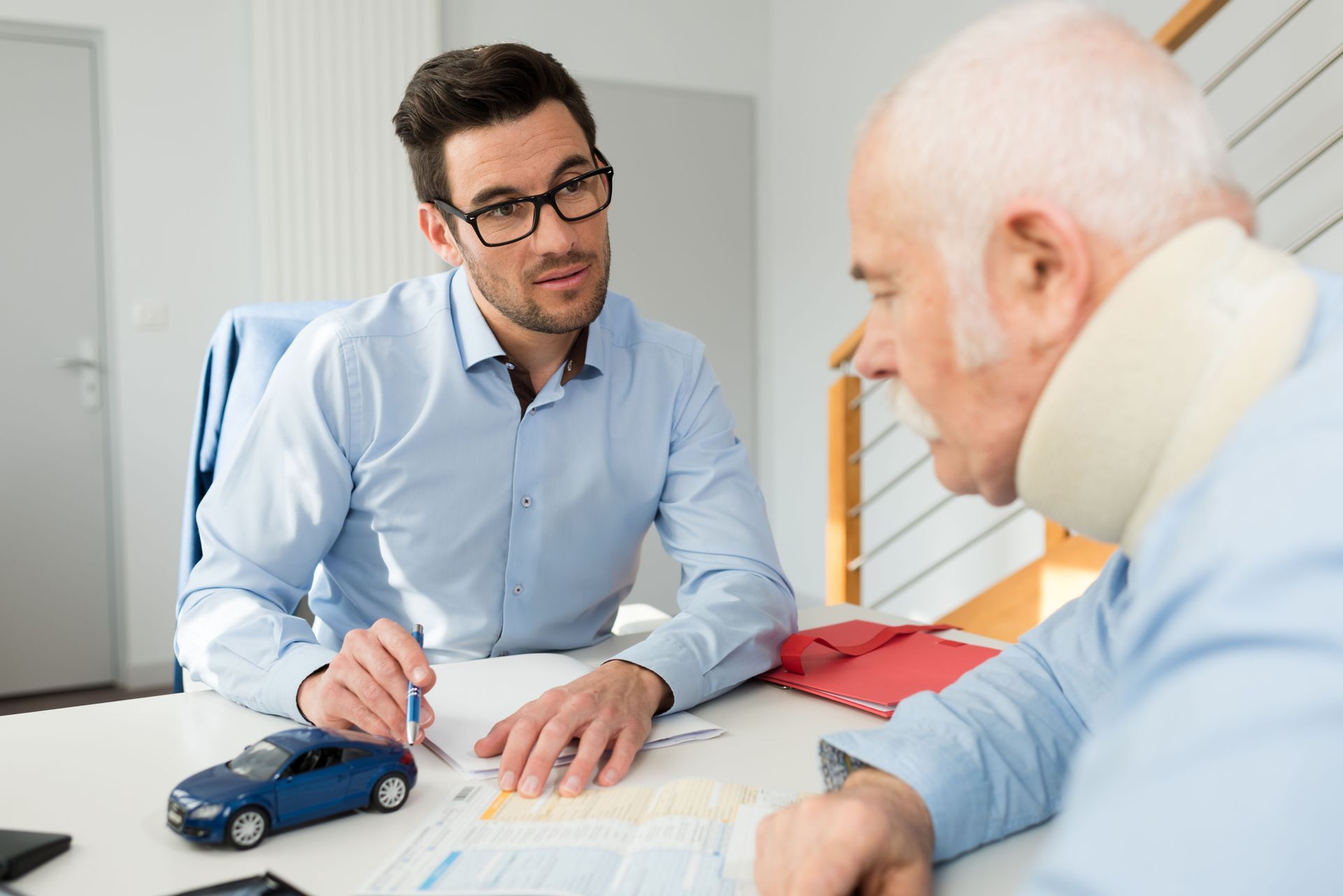 A person in a blue shirt consults with a patient wearing a neck brace at a table with a small blue toy car.
