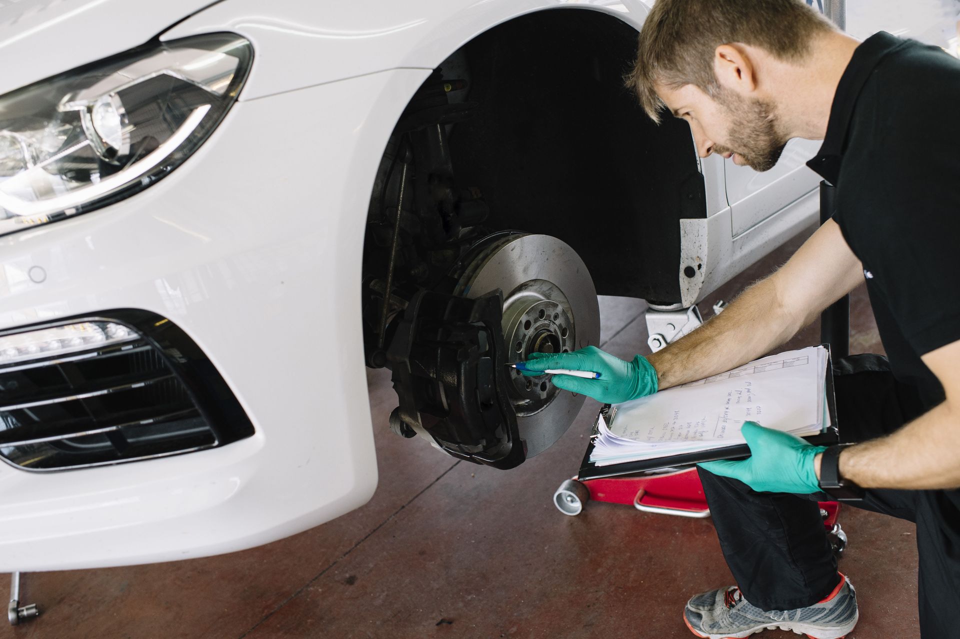 Auto technician inspecting a car brake system and taking notes on a clipboard in a repair shop.