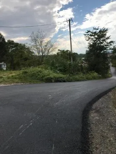 Asphalt road curves into a grassy area with trees, utility pole, and cloudy sky.