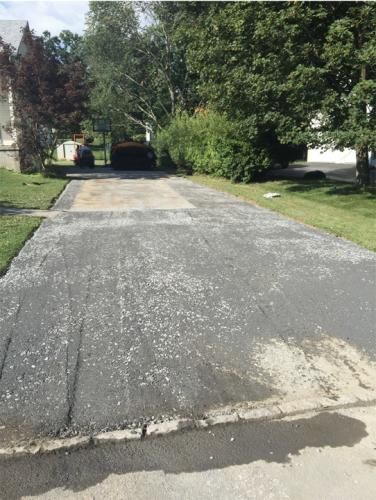 Asphalt driveway with scattered gravel, next to a grassy yard and trees.
