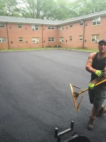 Man with rake stands on newly paved asphalt; apartment building in background.